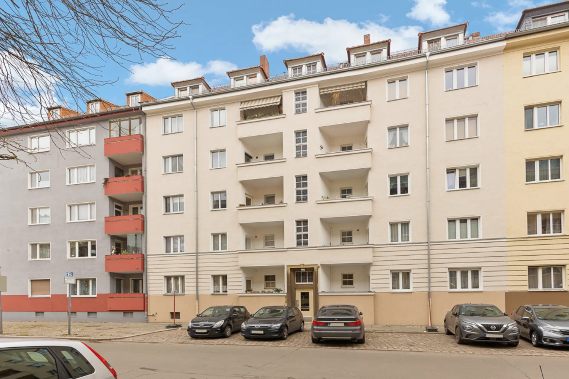 Mehrstöckiges Wohnhaus mit Balkonen, geparkten Autos davor und blauem Himmel im Hintergrund.