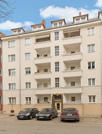 Mehrstöckiges Wohnhaus mit Balkonen, geparkten Autos davor und blauem Himmel im Hintergrund.