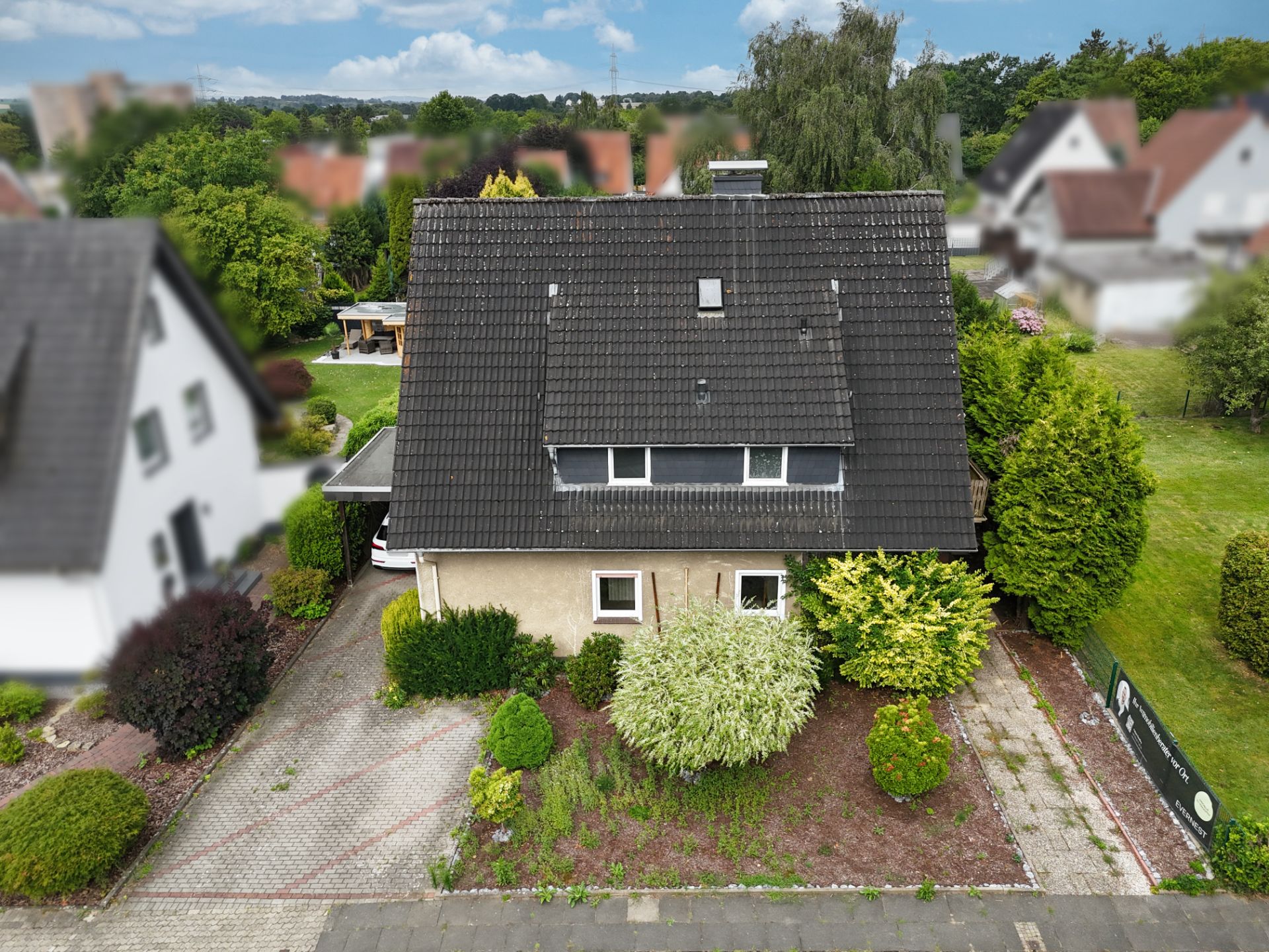 Zweistöckiges Haus mit dunklem Dach und Vorgarten mit verschiedenen Büschen und einer kleinen Einfahrt.