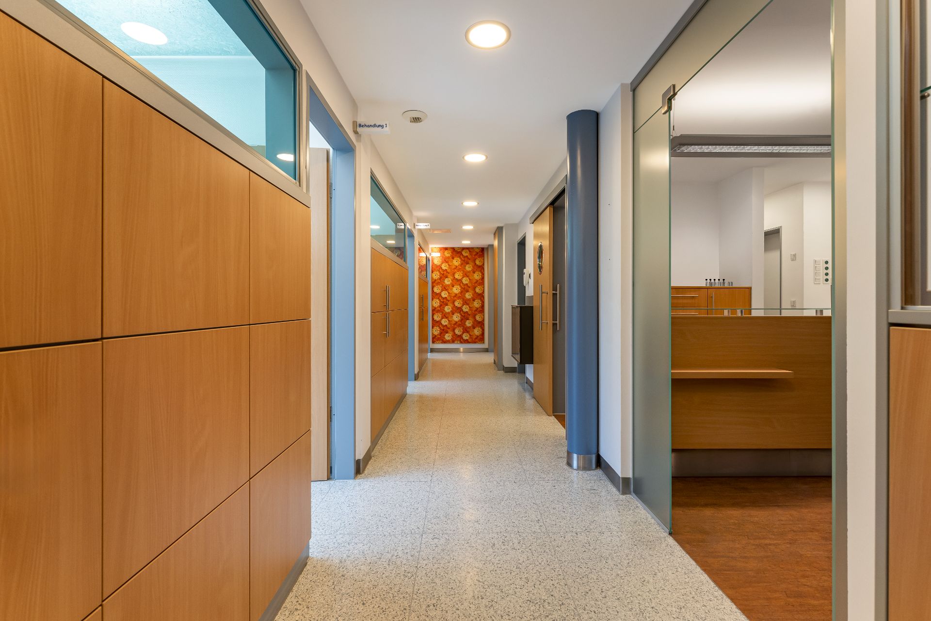 Heller Flur mit Holzwänden und Türen, sichtbarem Büro und bunter Blumenwand am Ende.