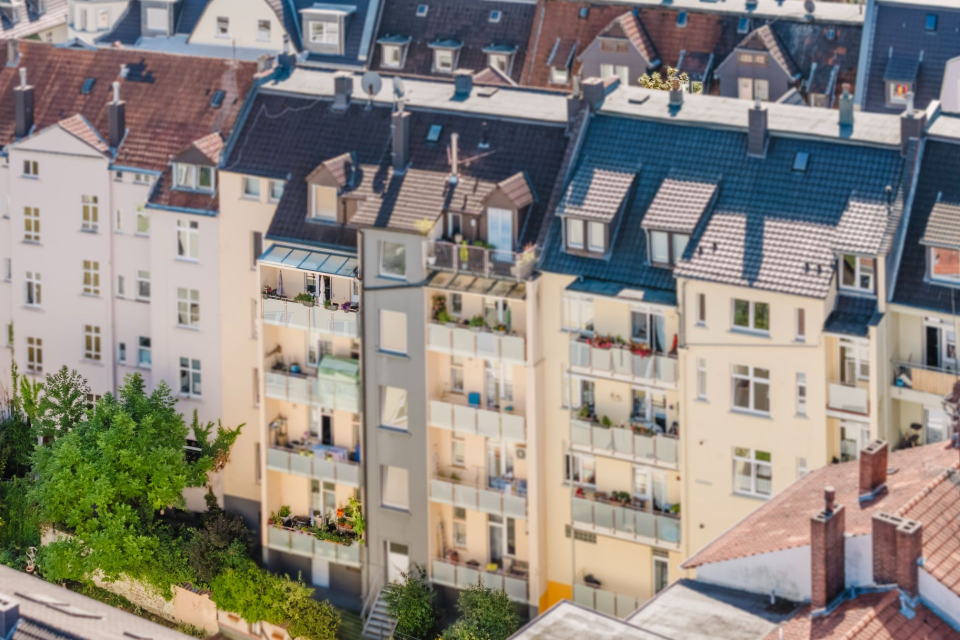 Mehrstöckiges Wohnhaus mit Balkonen und Pflanzen, davor Bäume und ein Gehweg.