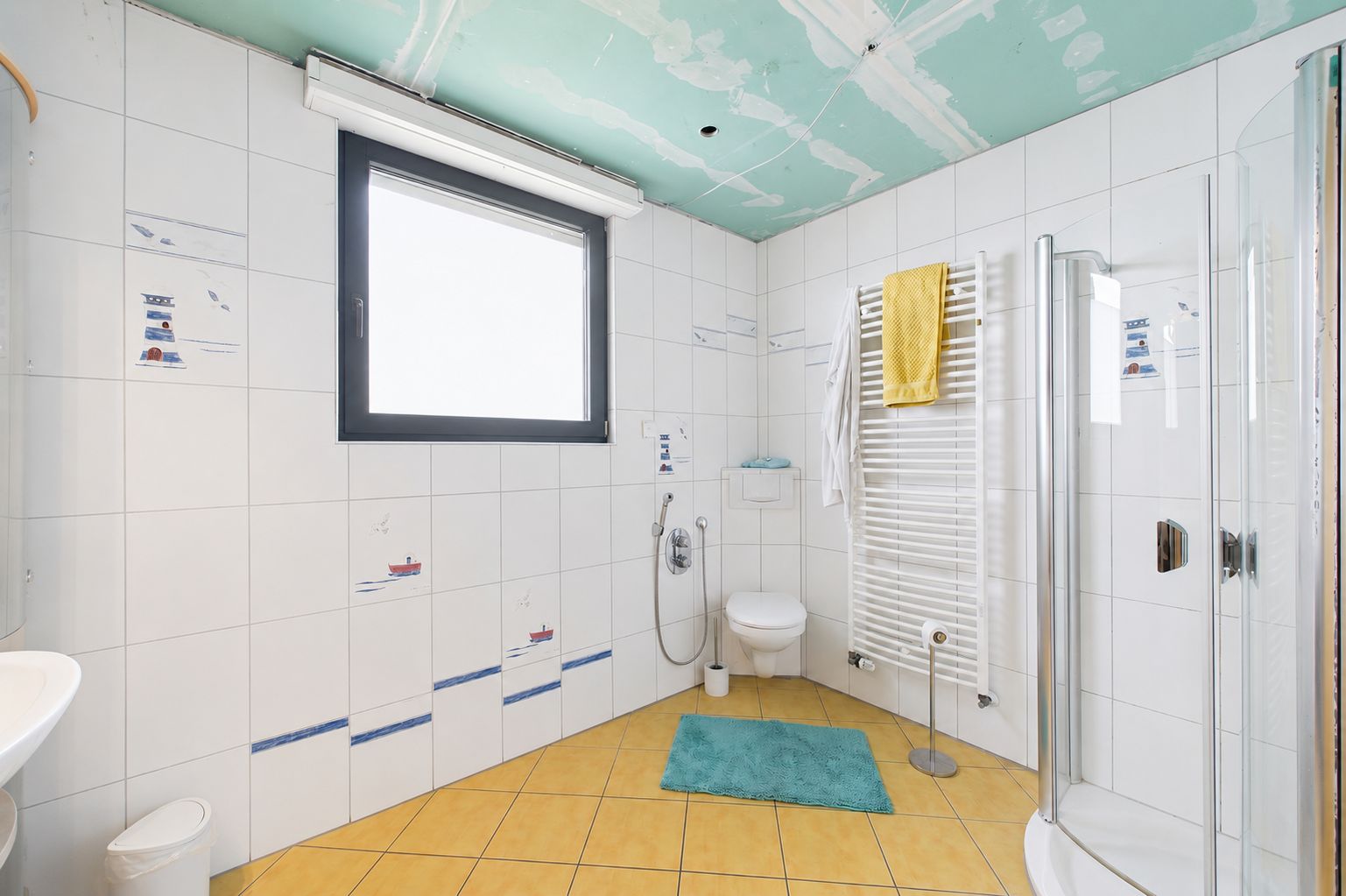 Helles Badezimmer mit Fenster, Dusche, Toilette, Handtuchheizkörper und türkisfarbener Matte.