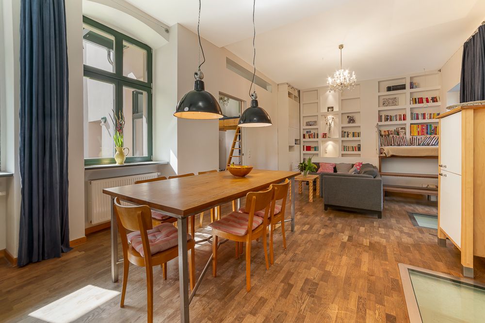 Helles Wohnzimmer mit Esstisch, vier Stühlen, Bücherregal, Sofa und großen Fenstern.
