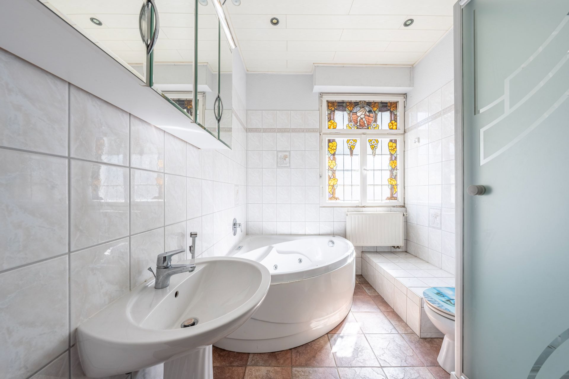 Helles Badezimmer mit Eckbadewanne, Waschbecken und verzierten Fenster.