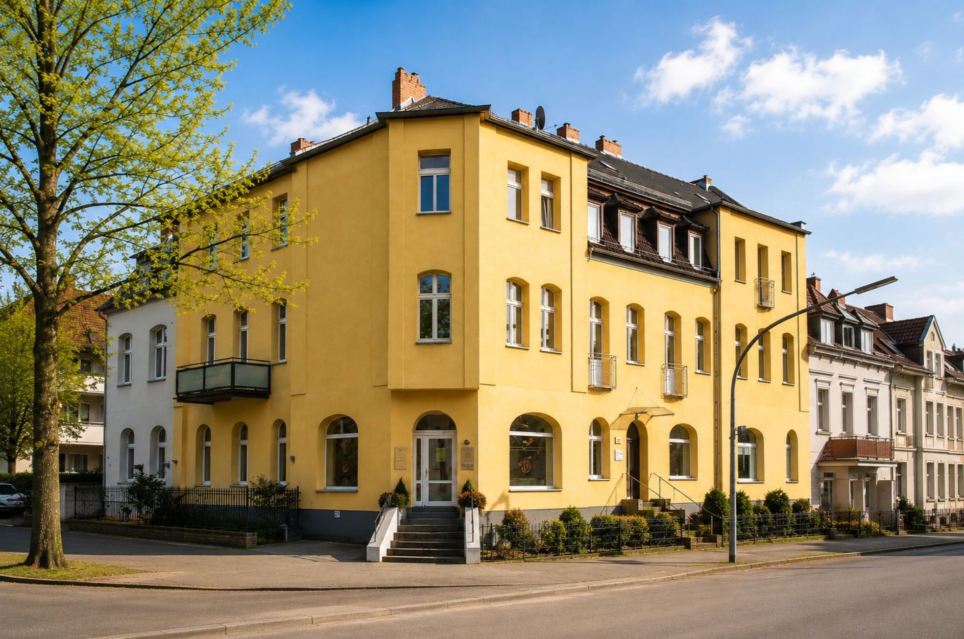 Dreistöckiges gelbes Wohnhaus mit mehreren Balkonen an einer Straßenecke.