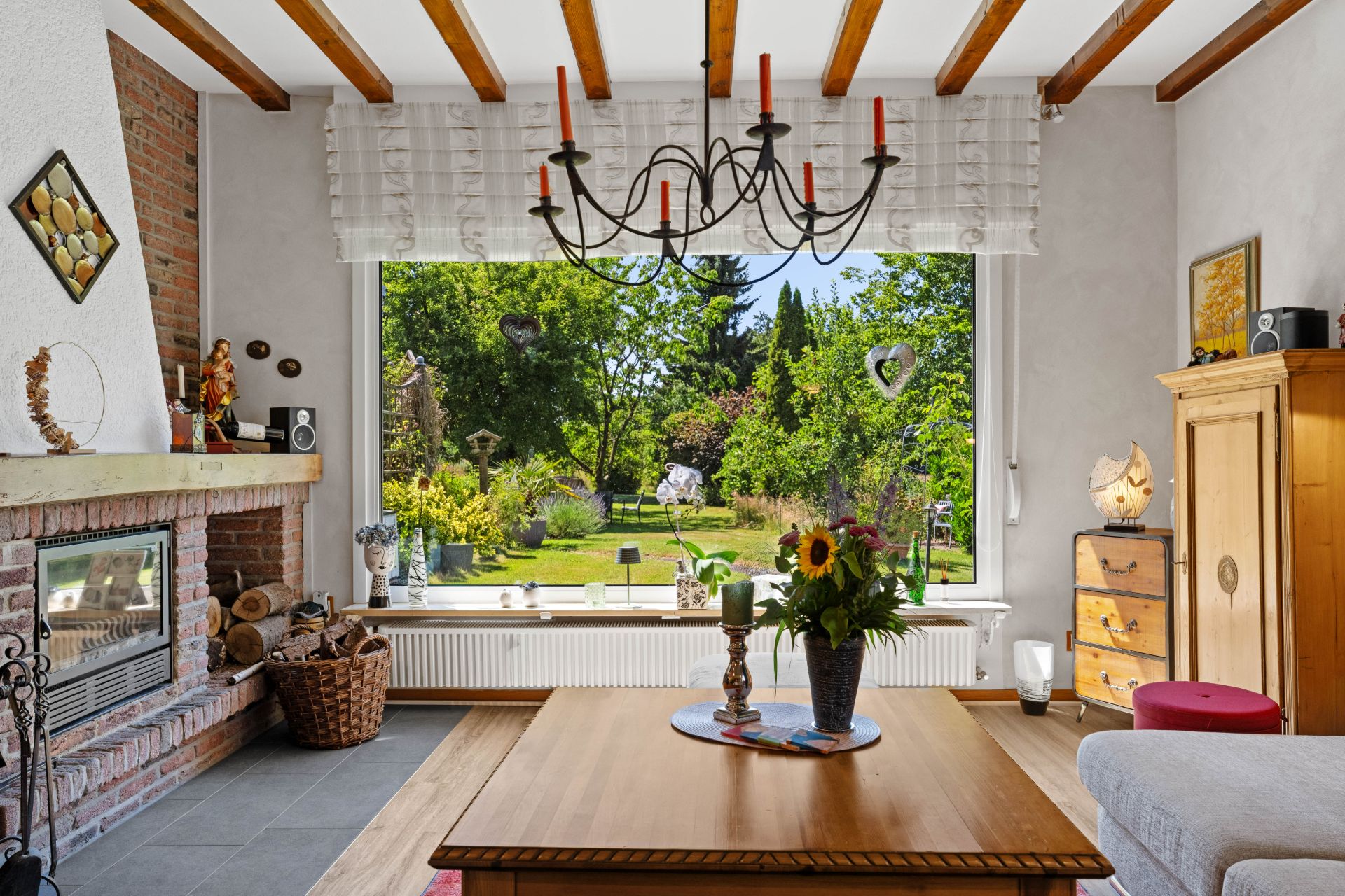 Helles Wohnzimmer mit Kamin, Holztisch, großem Fenster und Blick in einen grünen Garten.
