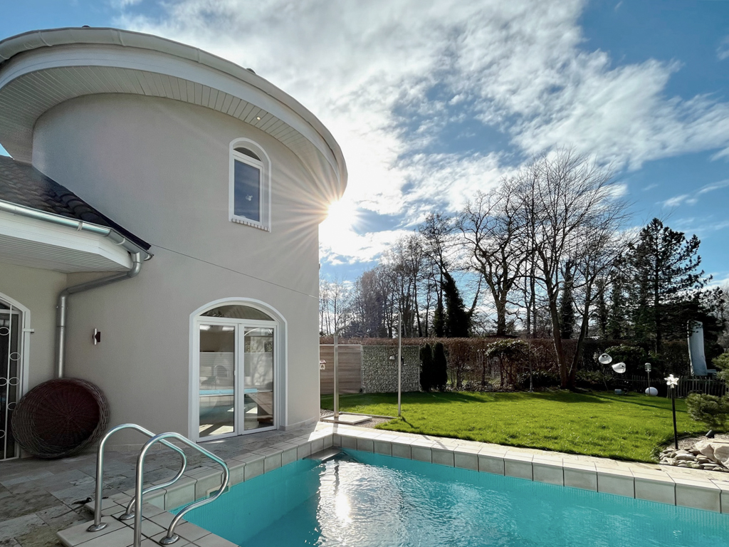 Haus mit rundem Turm, Pool im Garten, grüne Wiese und Bäume bei Sonnenschein.