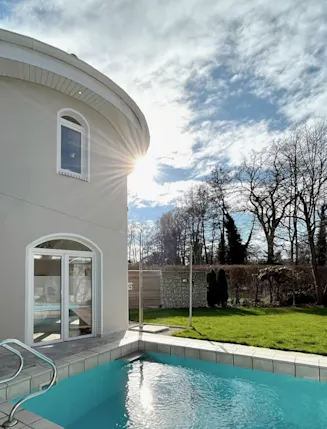 Haus mit rundem Turm, Pool im Garten, grüne Wiese und Bäume bei Sonnenschein.