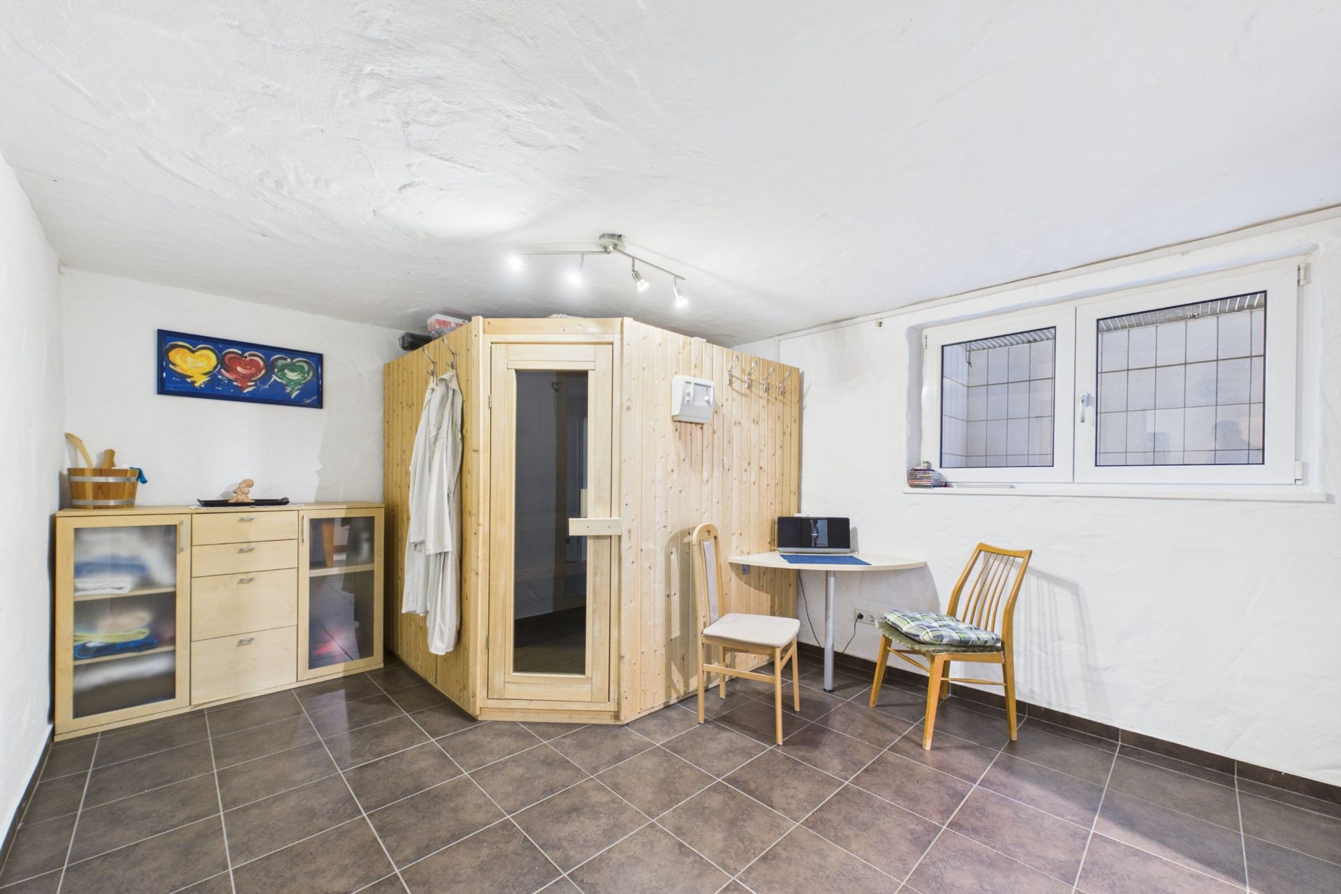 Raum mit Holzkabine, Schrank, Tisch, zwei Stühlen und Fenster auf gefliestem Boden.