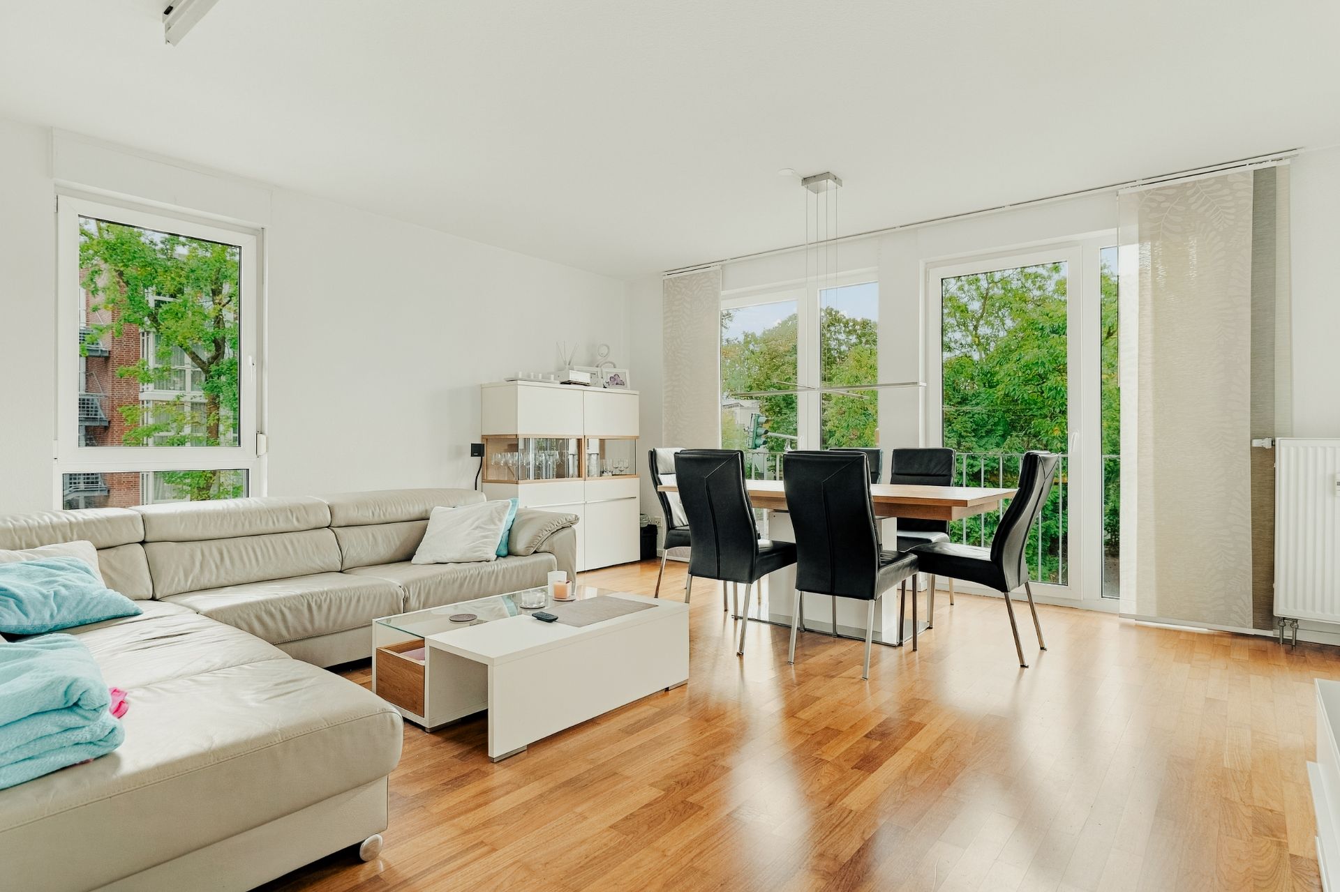 Helles Wohnzimmer mit Ecksofa, Esstisch und großen Fenstern mit Blick ins Grüne.