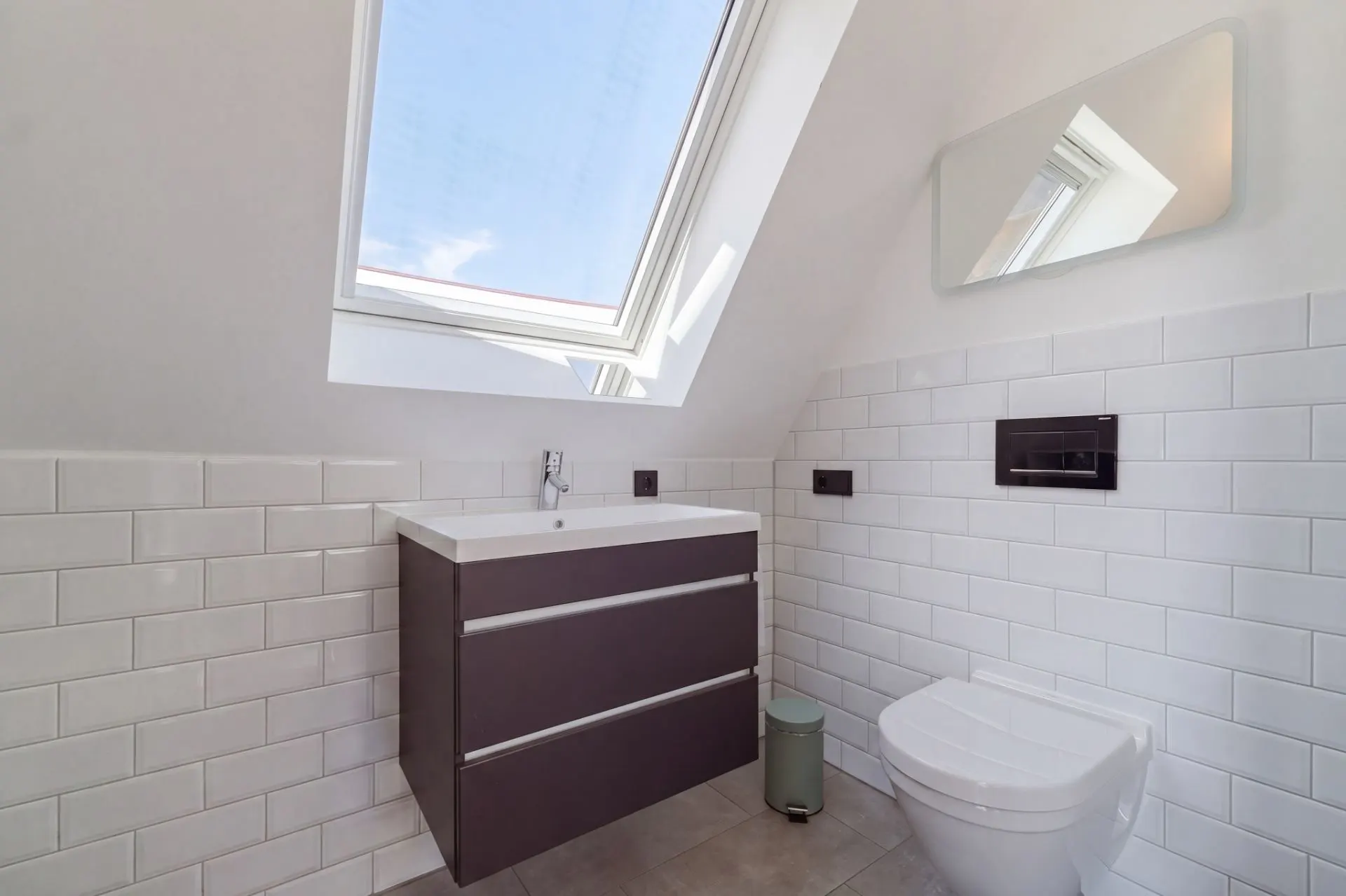 Modernes Bad mit Dachfenster, Waschbecken, Spiegel und Toilette, weiße Fliesen an der Wand.