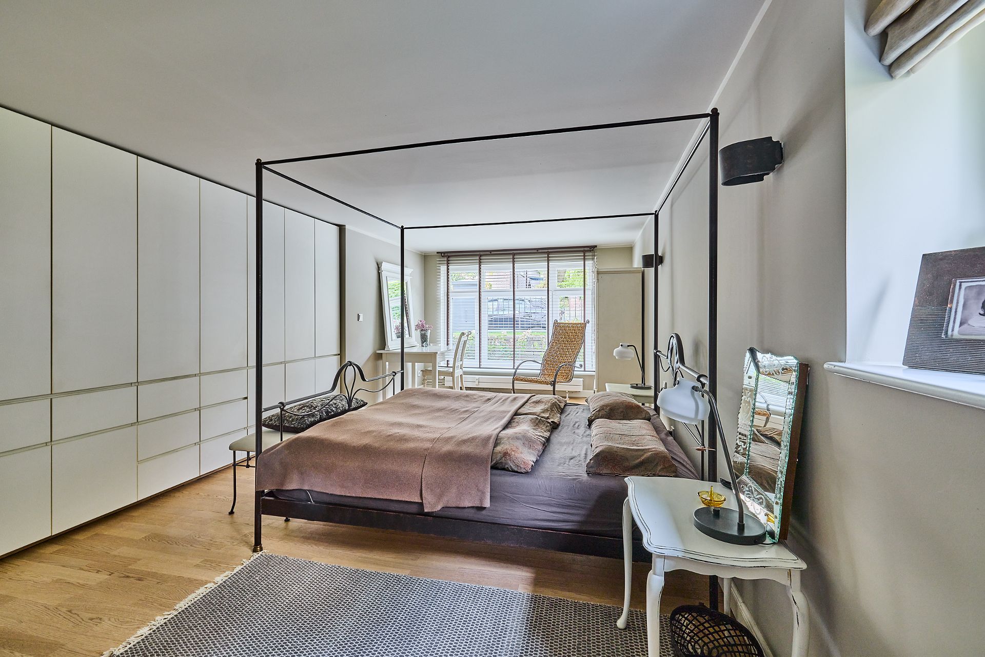 Modernes Schlafzimmer mit Himmelbett, großem Schrank, Schreibtisch und Sessel vor einem Fenster.