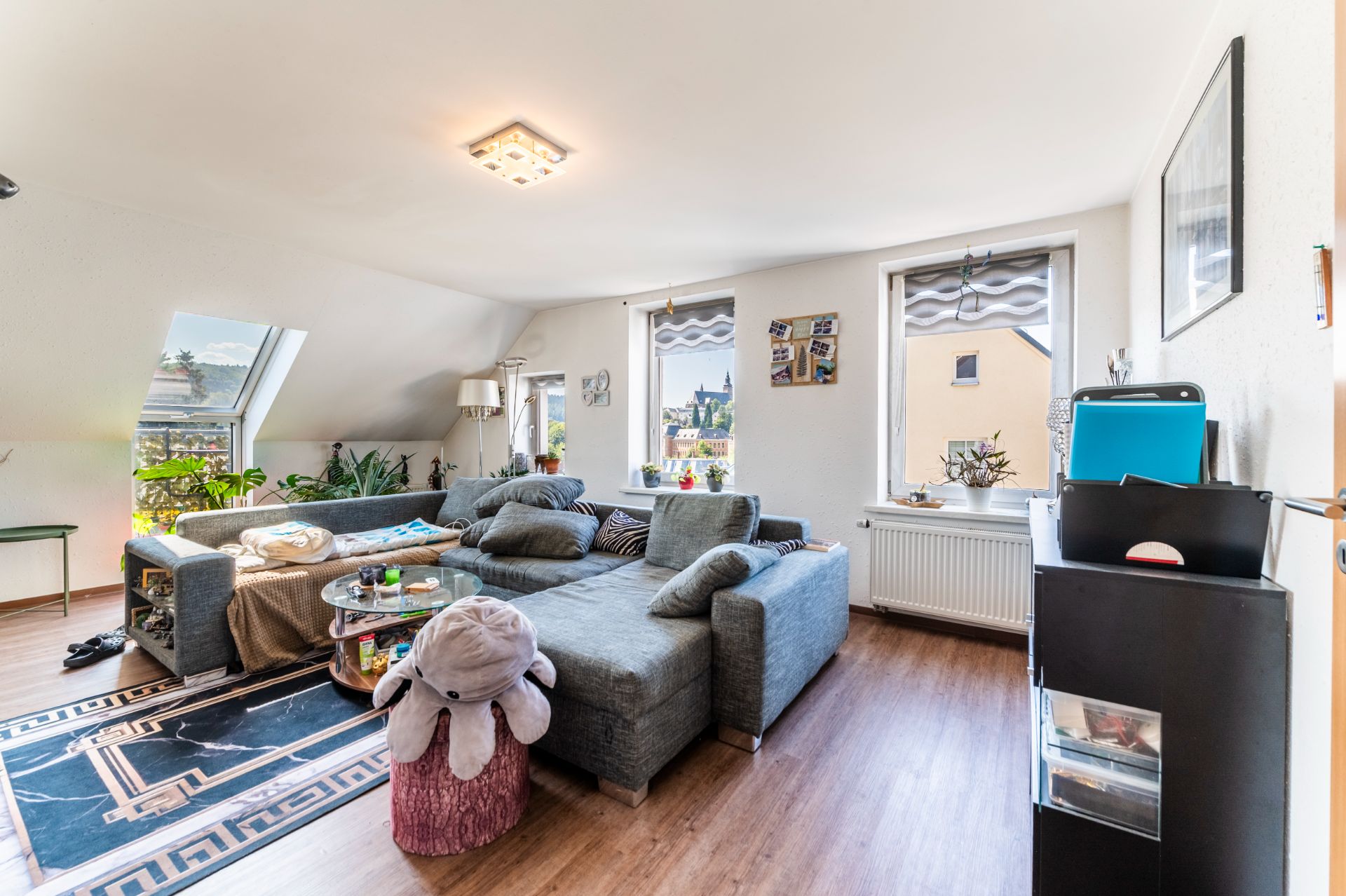 Helles Wohnzimmer mit Ecksofa, Holzboden, Pflanzen und großen Fenstern.