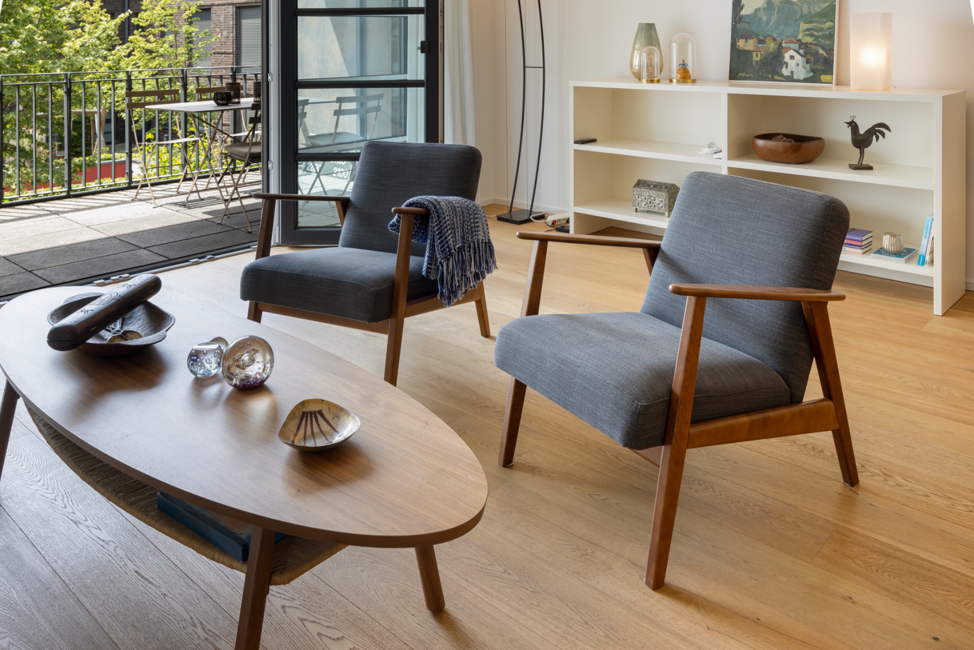 Helles Wohnzimmer mit zwei grauen Sesseln, Holztisch und Regal, Zugang zu Balkon mit Blick ins Grüne.