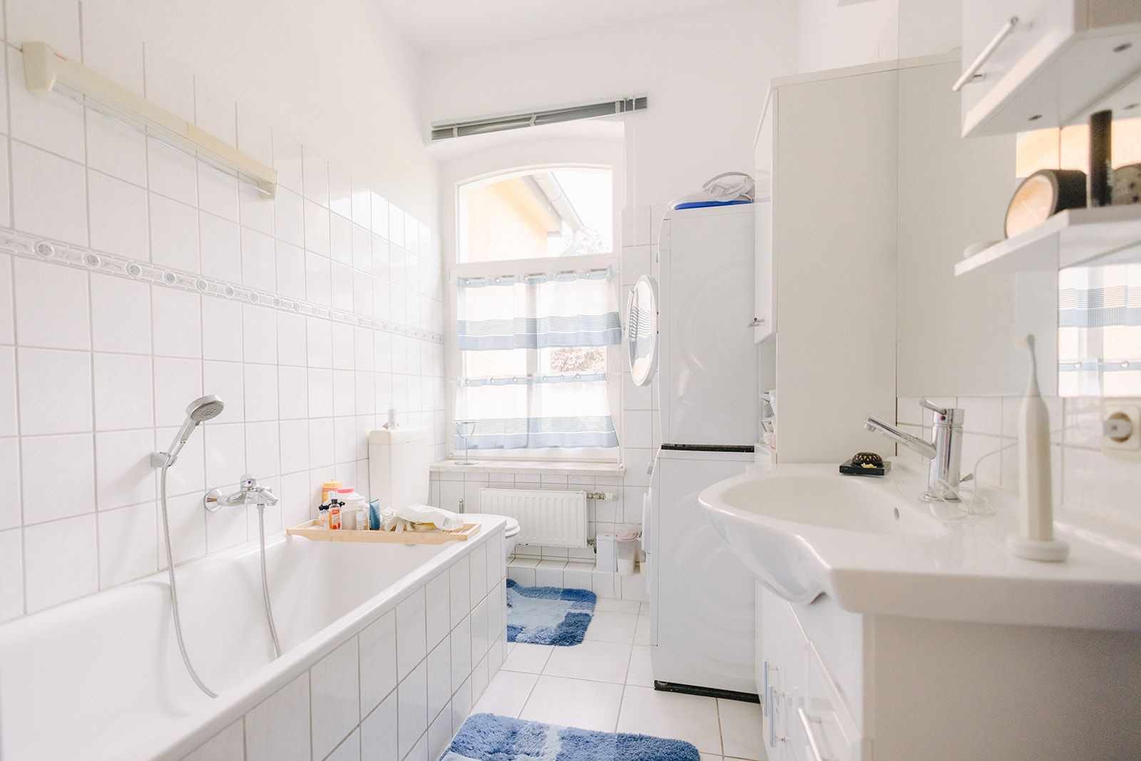 Helles Badezimmer mit Badewanne, Waschbecken, Waschmaschine und Fenster, blaue Badteppiche auf dem Boden.