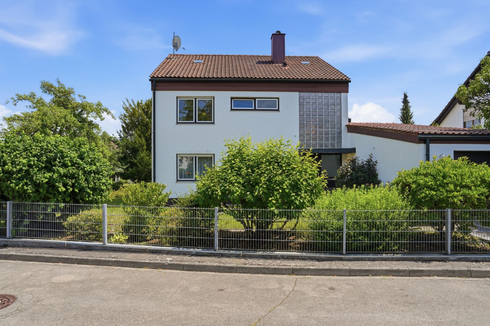 Zweistöckiges Wohnhaus mit Satteldach, großem Fensterblock und Garten, umgeben von einem Metallzaun.