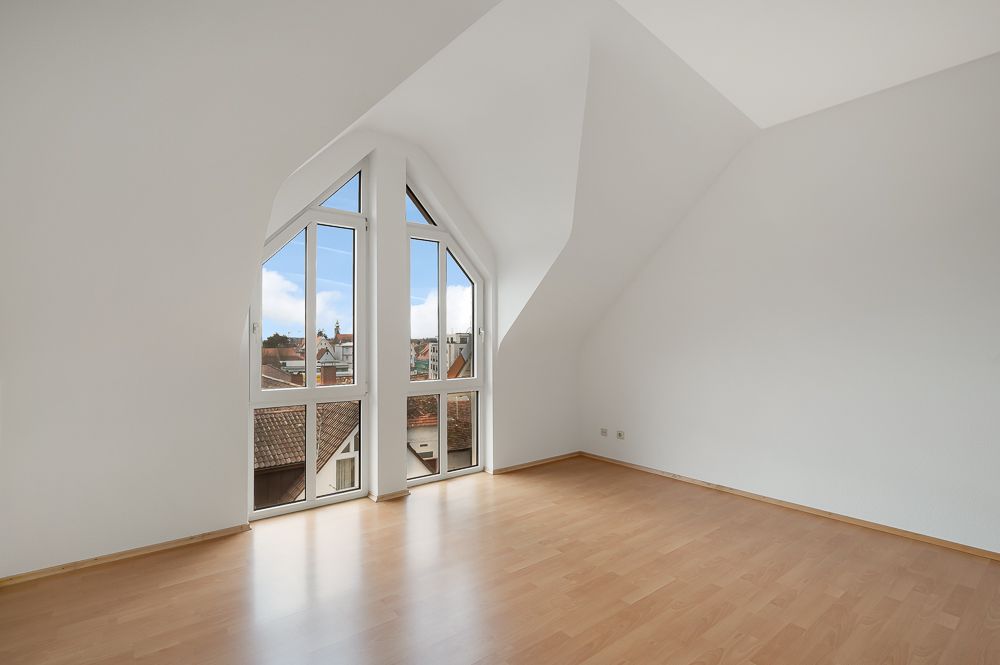 Helles Zimmer mit schrägen Wänden, großem Fenster und Parkettboden.