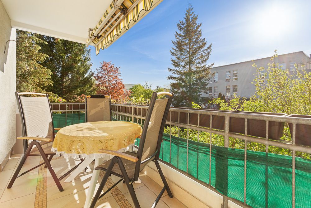 Heller Balkon mit Tisch, vier Stühlen und Ausblick auf Bäume und ein Wohngebäude.