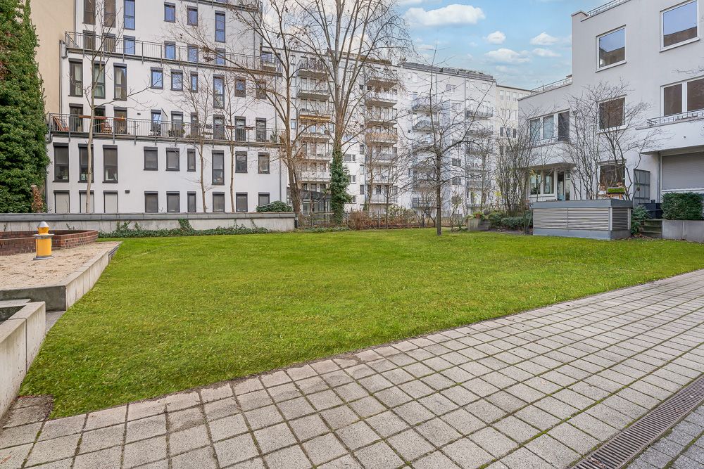Grüne Rasenfläche mit Gehweg, umgeben von mehrstöckigen Wohngebäuden und einigen Bäumen.
