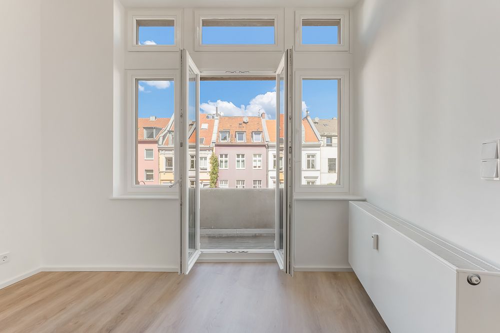 Helles Zimmer mit Holzboden, bodentiefen Fenstern und Blick auf Nachbarhäuser.
