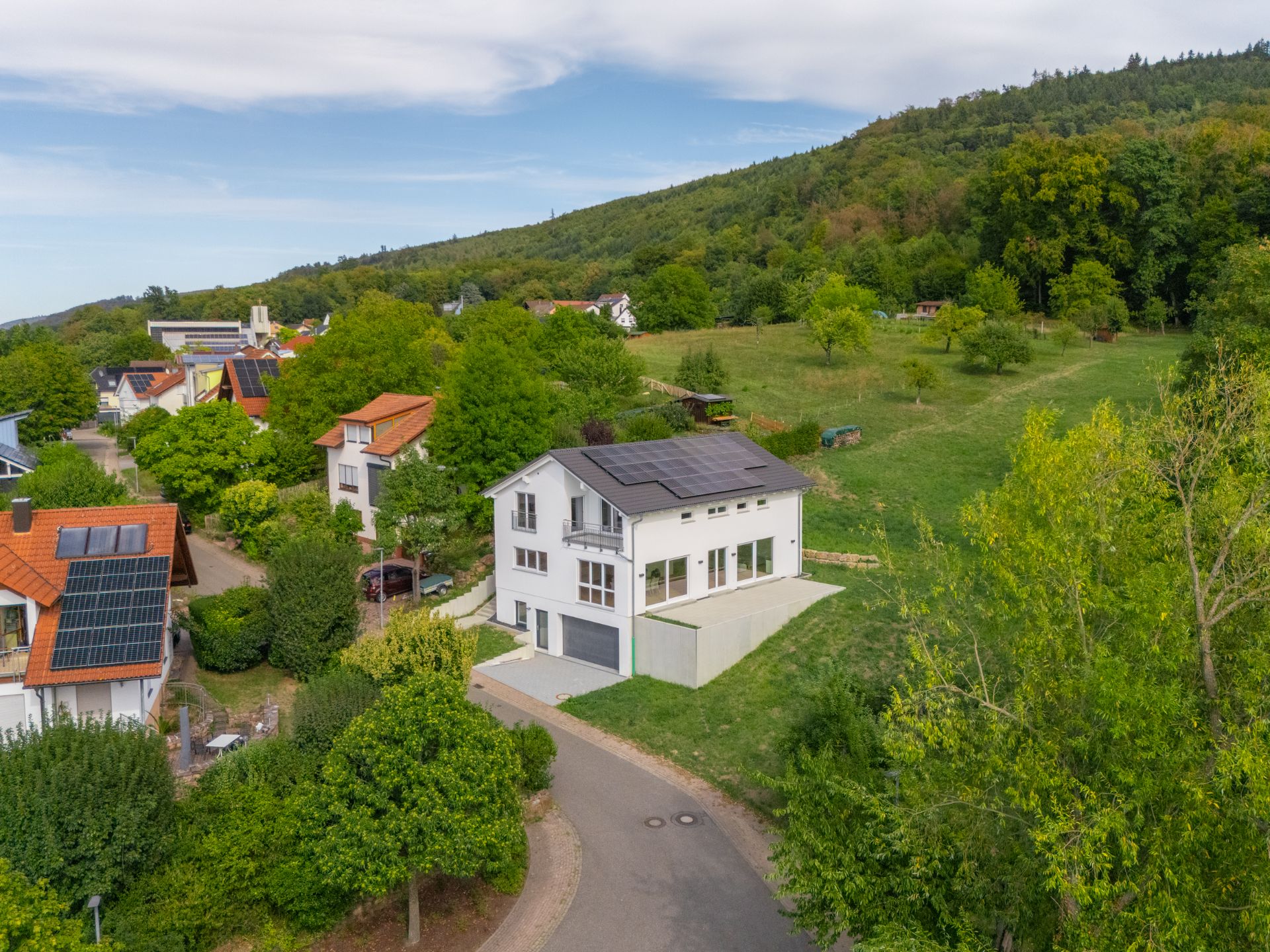 Modernes weißes Haus am Waldrand mit Solarpanels und großem Garten, umgeben von grünen Bäumen.