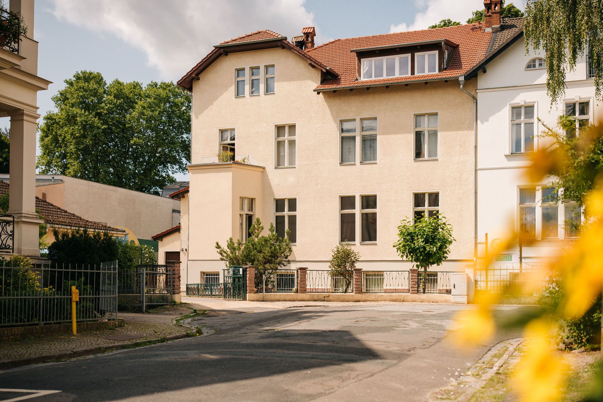 Mehrstöckiges Wohnhaus mit gelber Fassade und rotem Dach an einer ruhigen Straße.