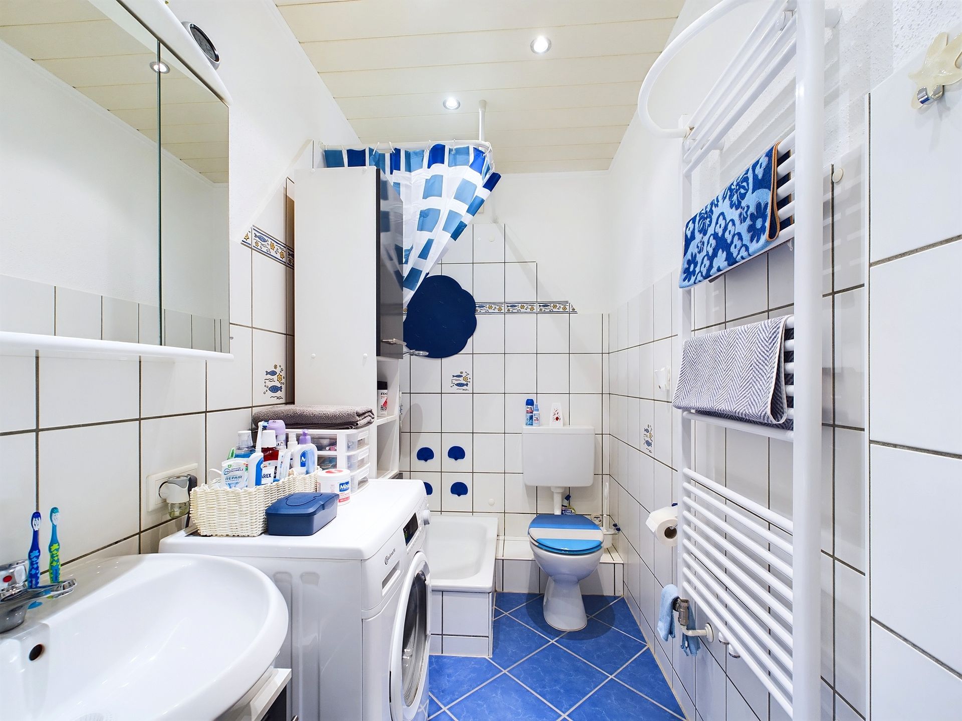 Kleines Badezimmer mit blauen Fliesen, Waschmaschine, Dusche, Toilette und Handtuchheizkörper.