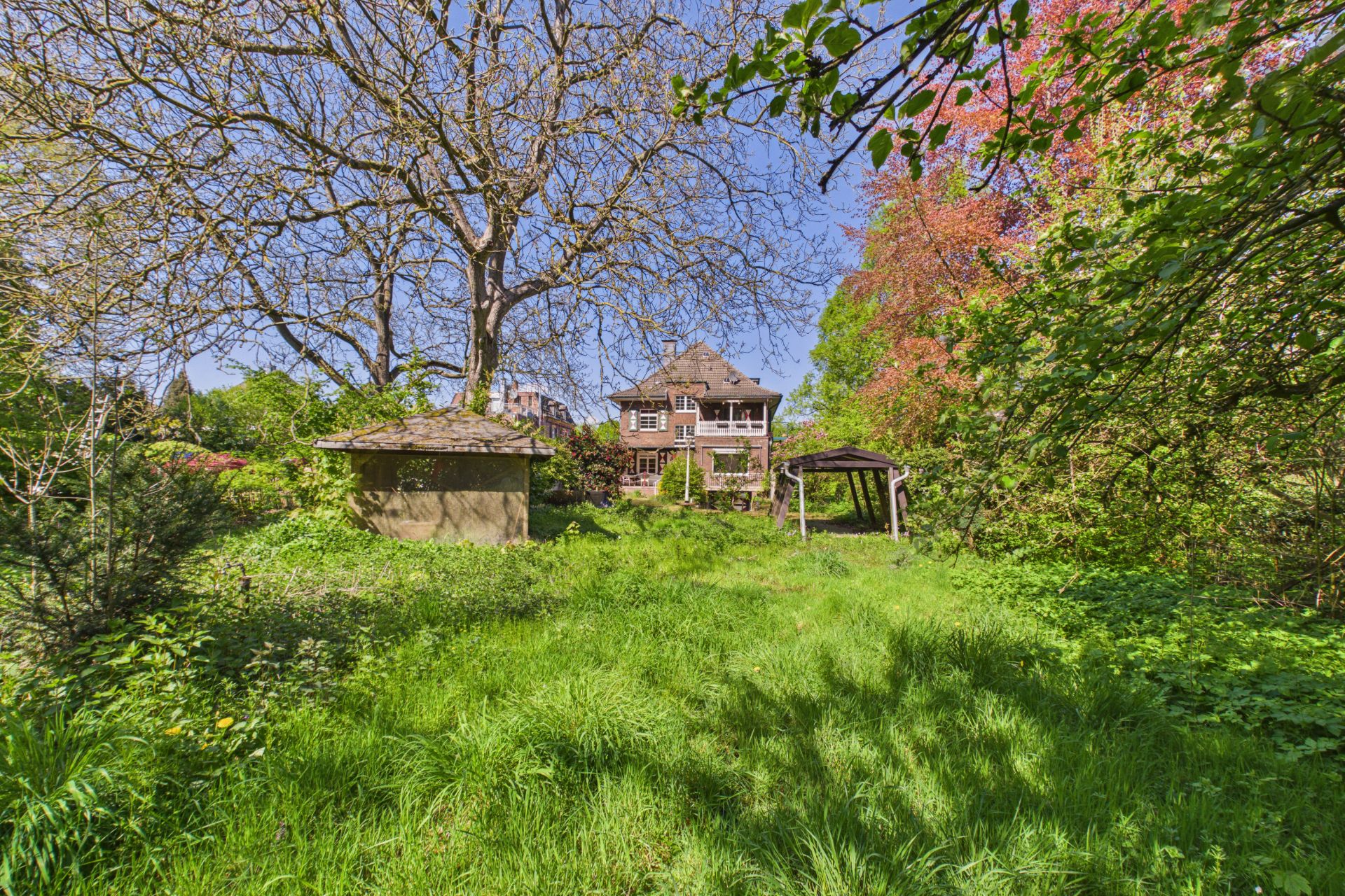 Großer Garten mit altem Haus, Bäumen, Gras, Schuppen und Holzpavillon bei Sonnenschein.