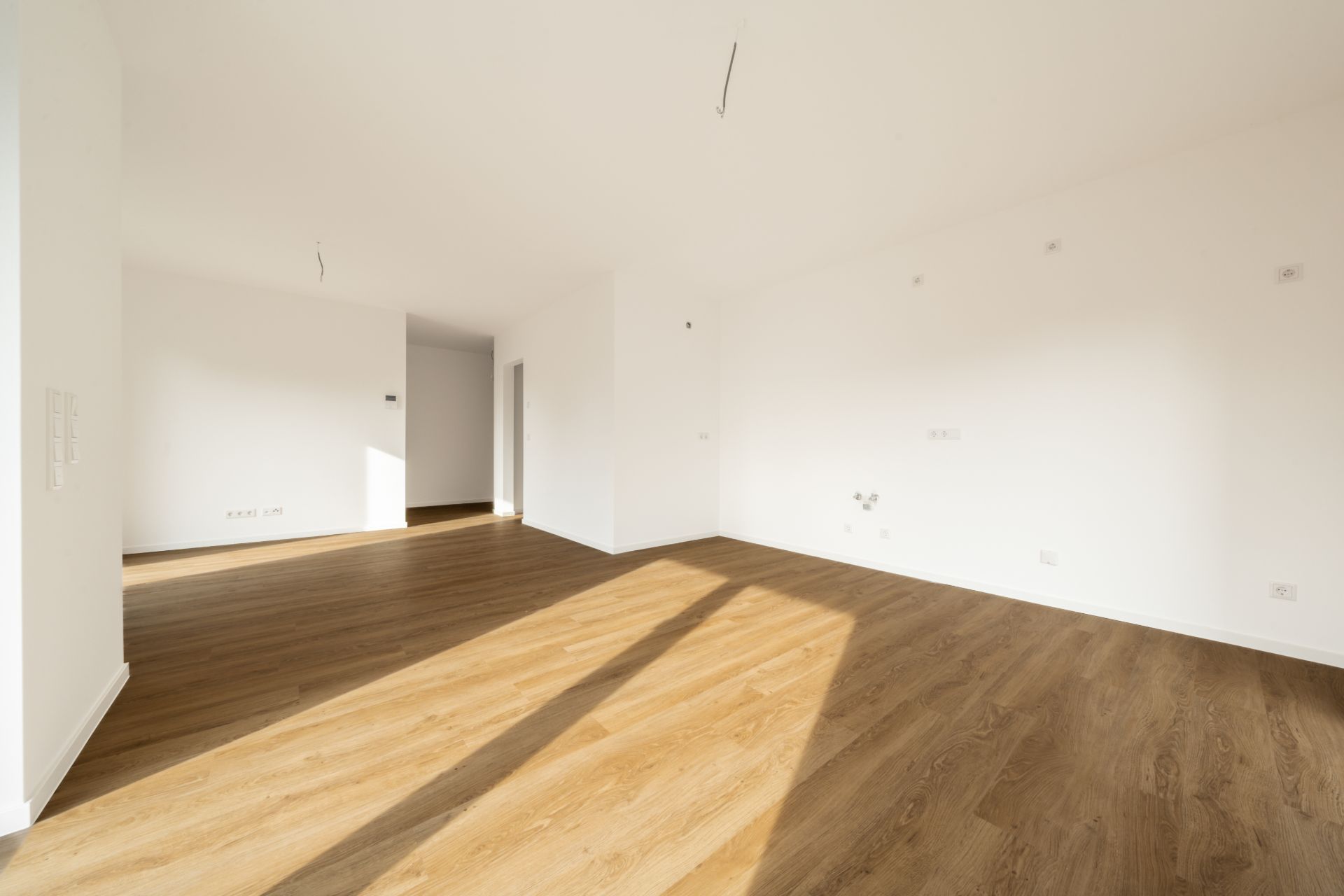 Heller, leerer Raum mit Holzboden und weißen Wänden, große Fenster lassen Sonnenlicht herein.