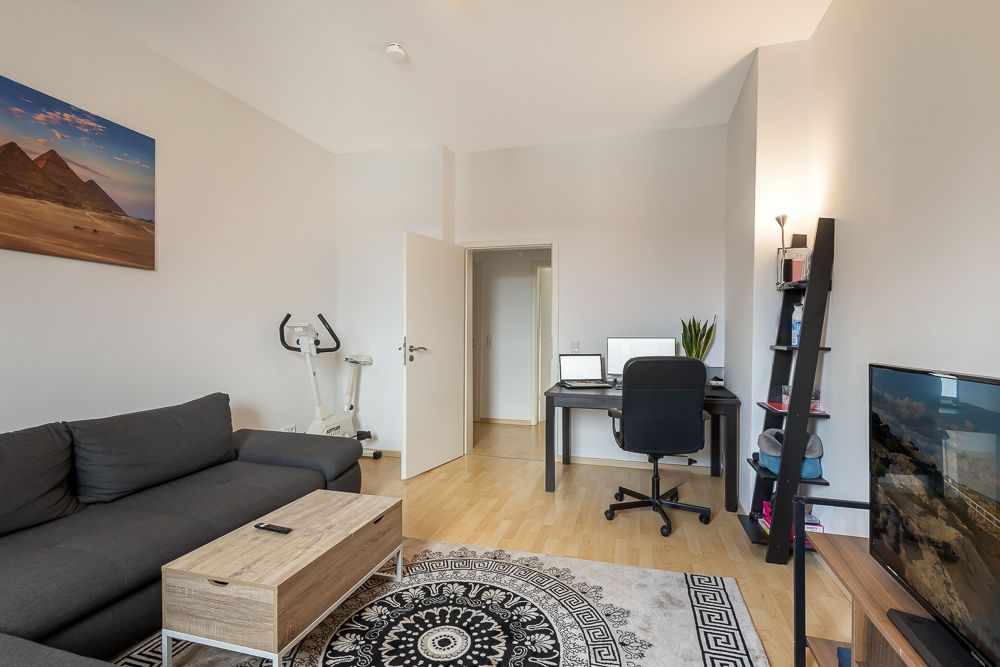 Wohnzimmer mit grauem Sofa, Schreibtisch, Fernseher, Teppich und einem Bild an der Wand.