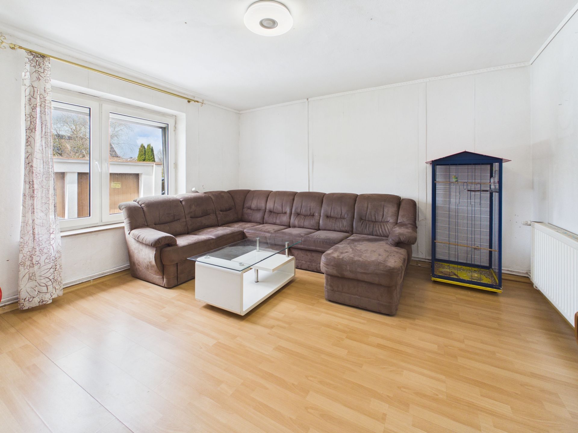 Wohnzimmer mit braunem Ecksofa, Glastisch und Vogelkäfig auf hellem Laminatboden.