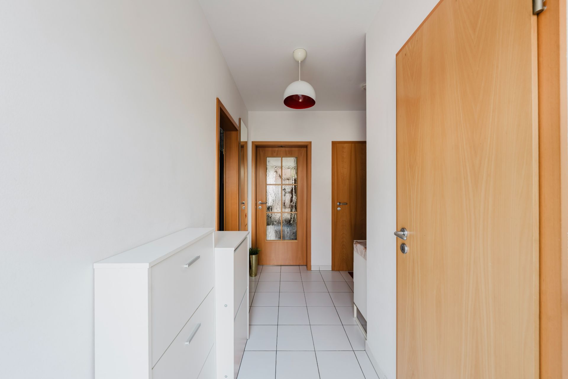 Heller Flur mit weißen Fliesen, Holztüren und weißem Schrank, Deckenlampe sichtbar.