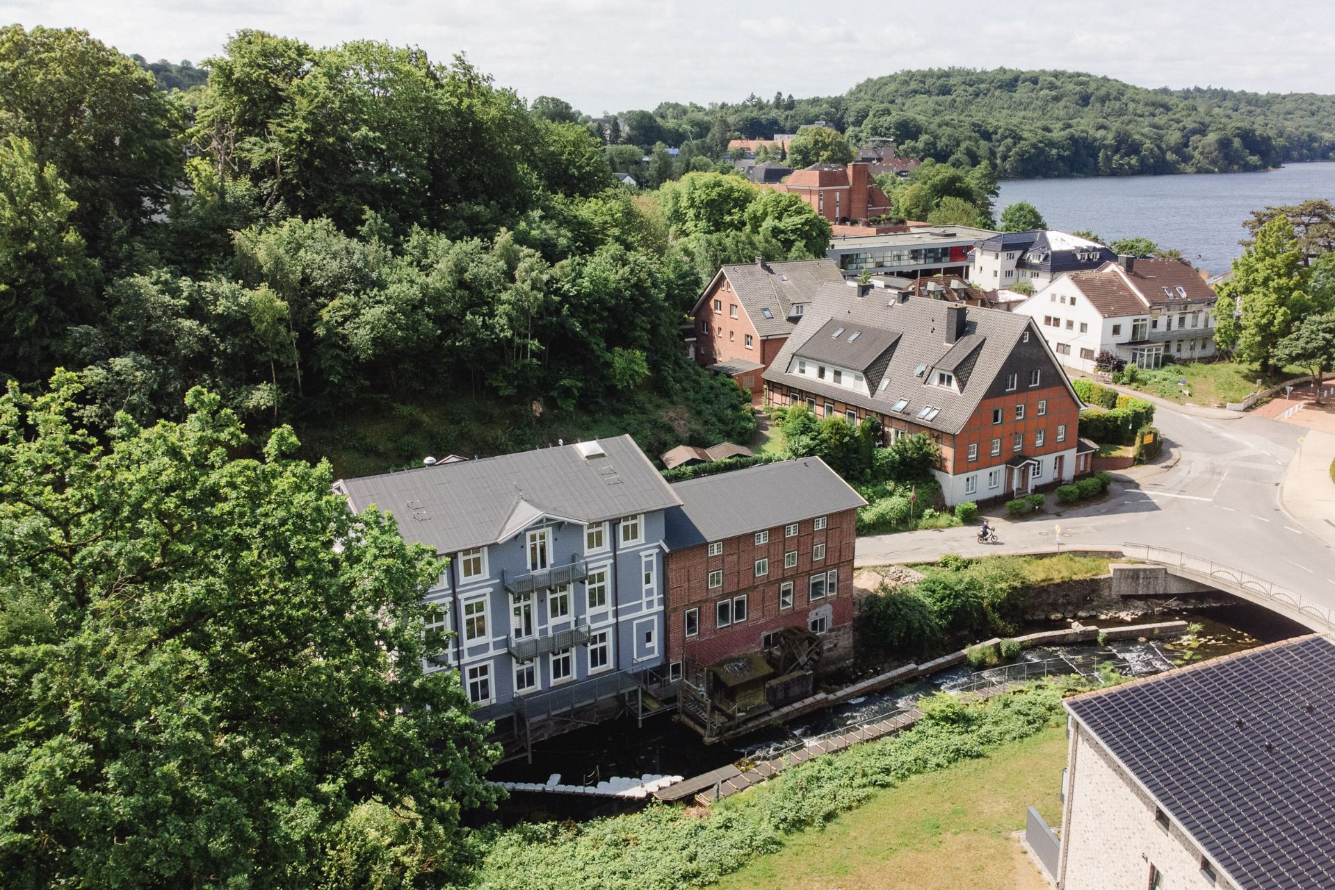 Wohnhäuser am Fluss, umgeben von Bäumen und Grünfläche, im Hintergrund ein See.