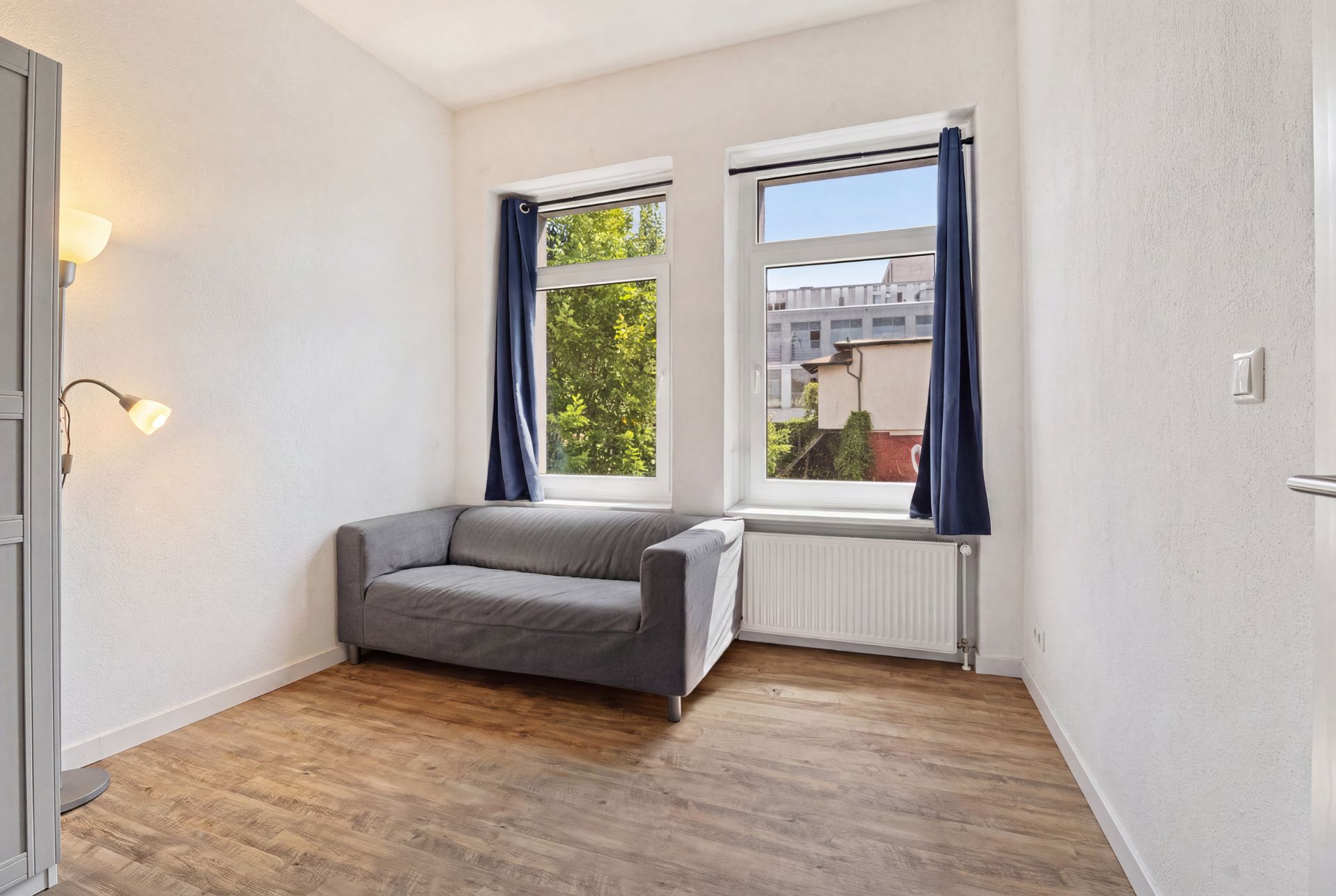 Helles Zimmer mit Holzfußboden, grauem Sofa und zwei Fenstern mit blauen Vorhängen.