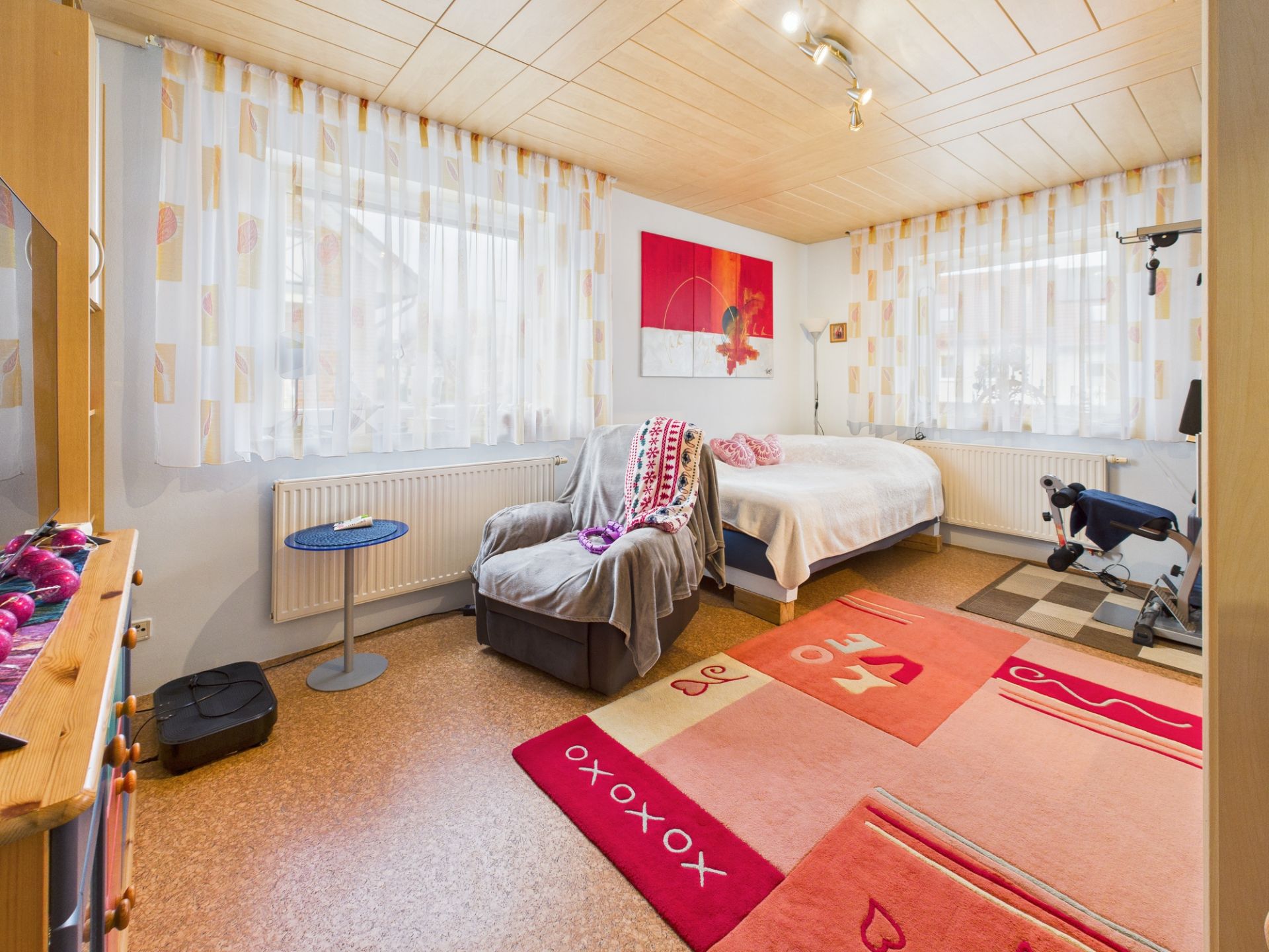 Helles Schlafzimmer mit Bett, Sessel, buntem Teppich und Fitnessgerät am Fenster.