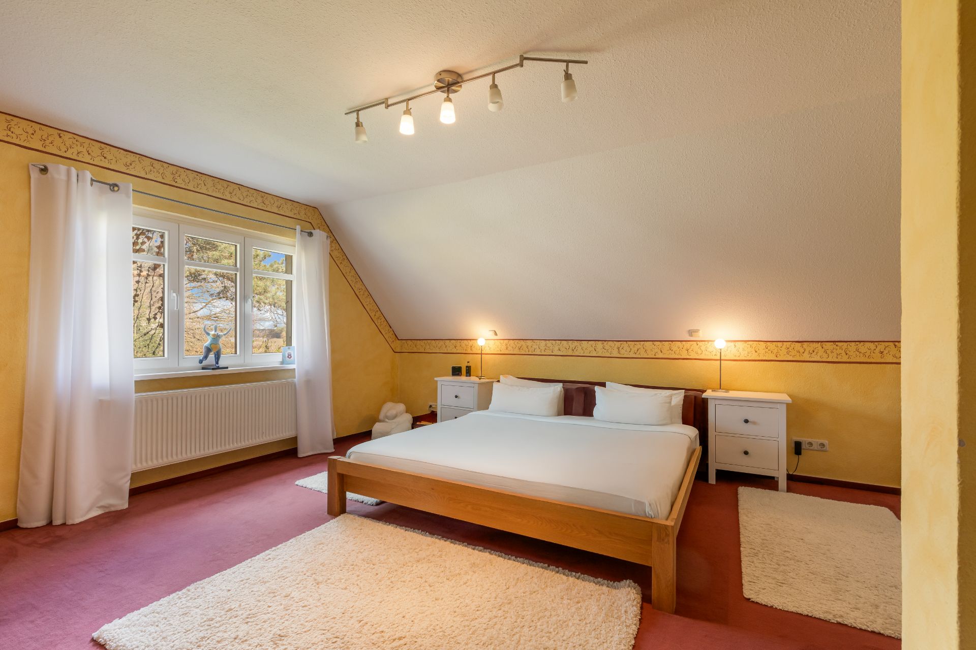 Helles Schlafzimmer mit Doppelbett, zwei Nachttischen, Teppichen und großem Fenster mit weißen Vorhängen.