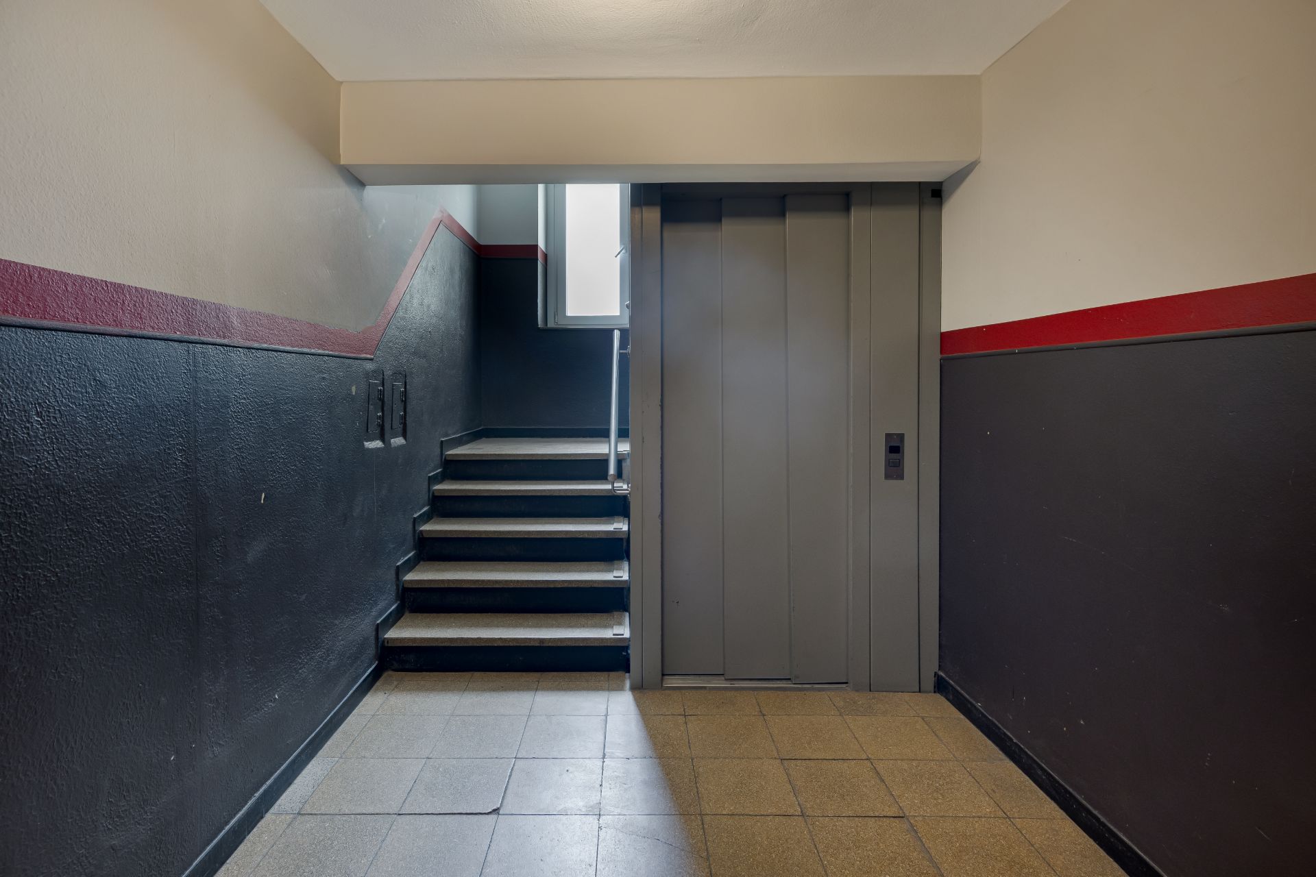 Treppenhaus mit Aufzug und Fenster, Wände dunkel gestrichen mit rotem Streifen.