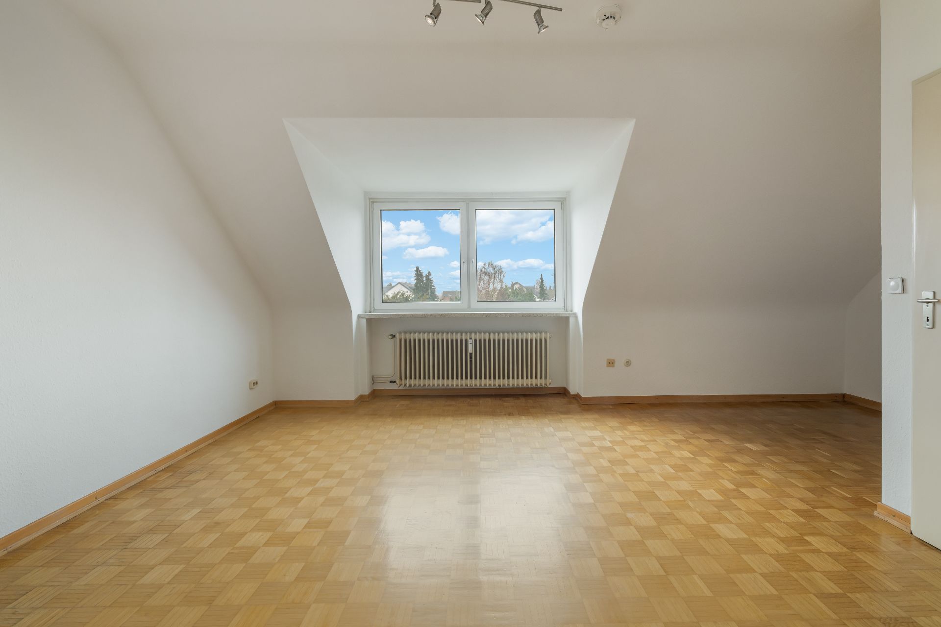 Leerer Raum mit Parkettboden, weißer Wand, großem Fenster und Heizkörper darunter.