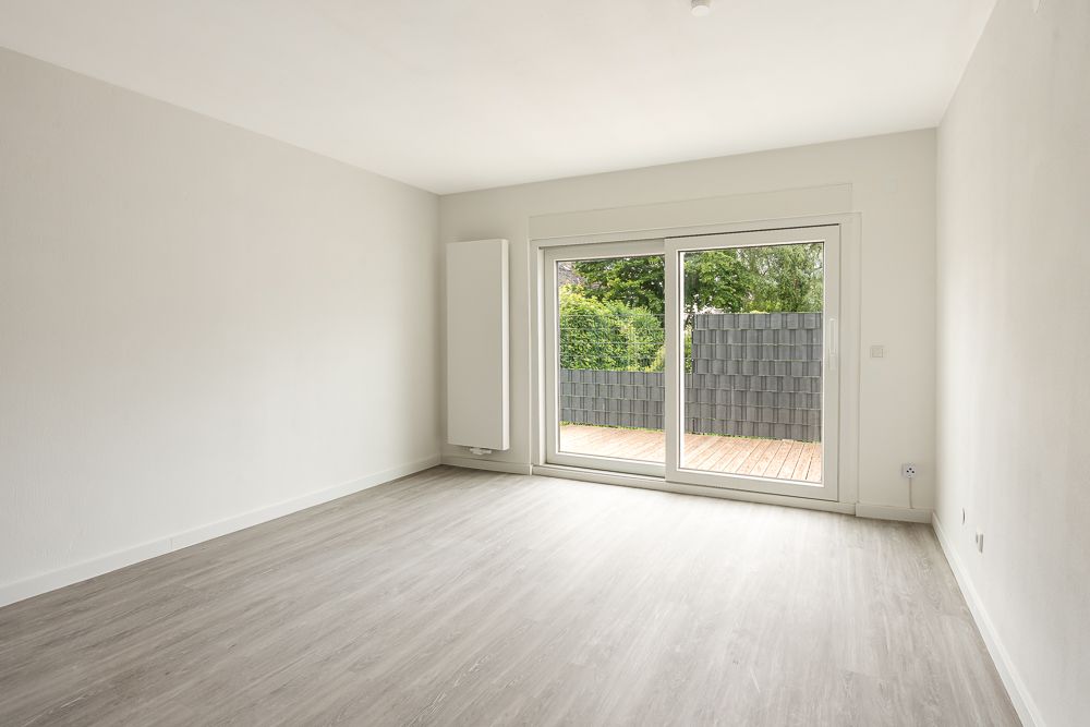 Leerer Raum mit hellen Wänden, großem Fenster und Blick auf Terrasse mit Holzboden.