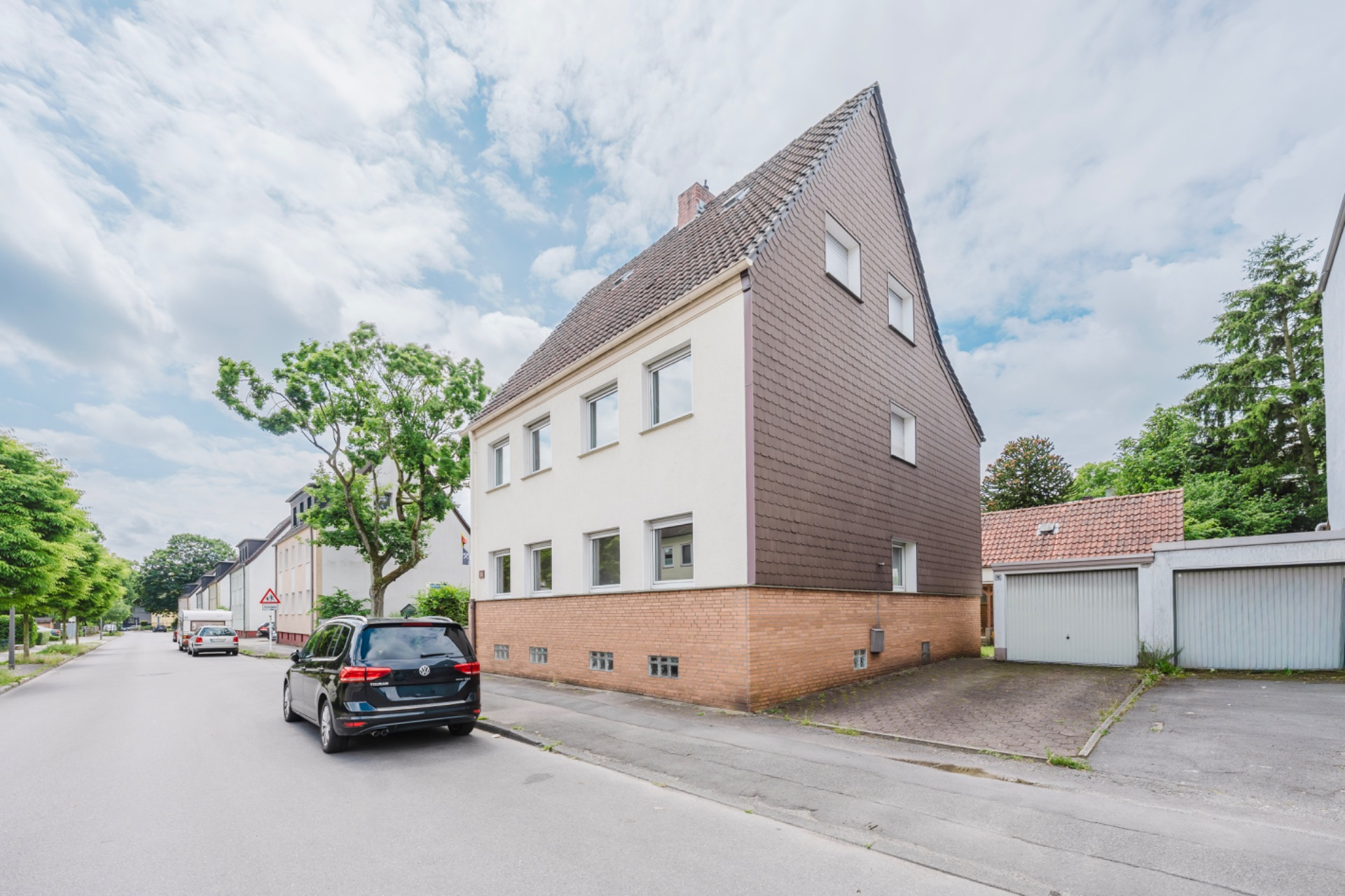 Eckhaus mit braunem Dach an einer ruhigen Wohnstraße, geparkte Autos und Garagen daneben.