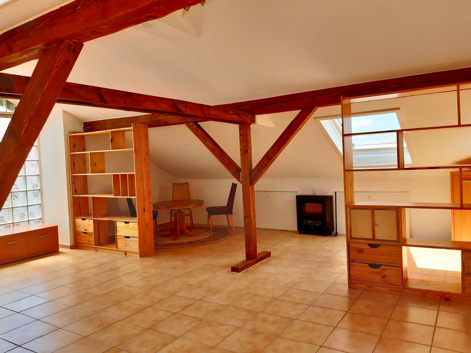 Helles Dachzimmer mit Holzbalken, Regalen, Tisch und Stühlen, Fliesenboden und Dachfenster.