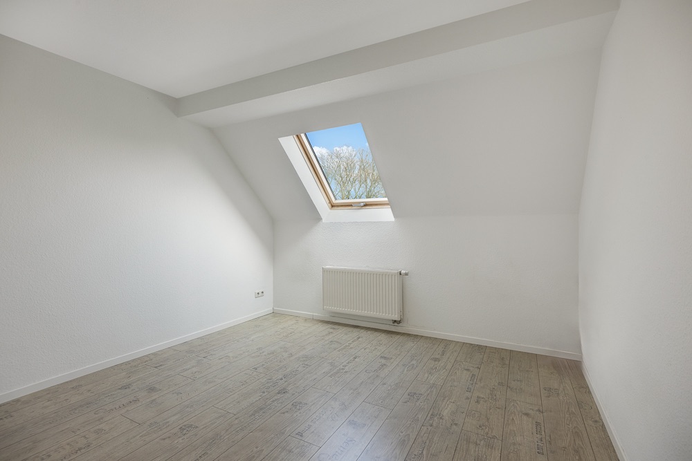Heller leerer Raum mit Dachschrägen, Holzboden, Heizkörper und Fenster im Dach.