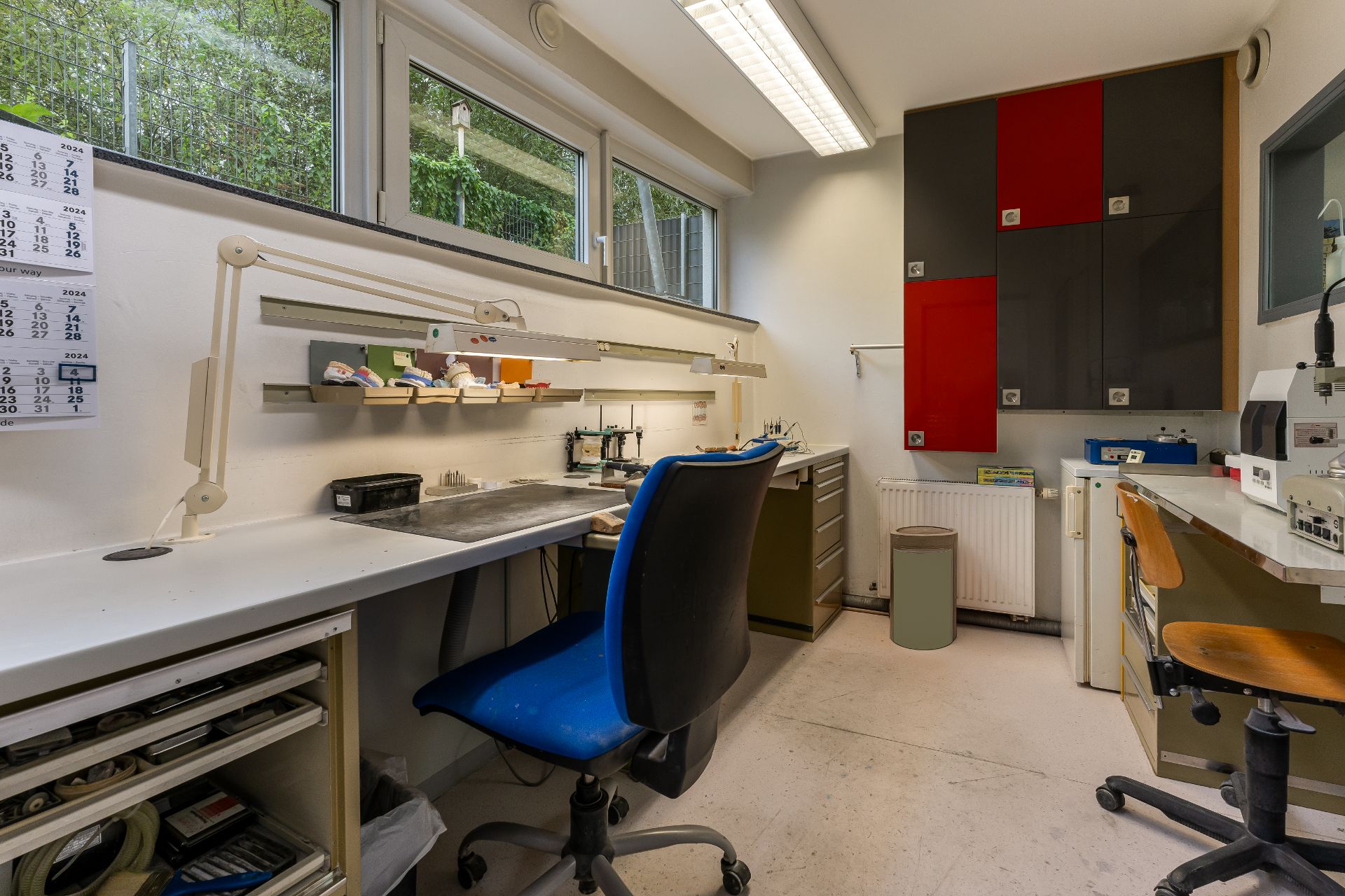 Arbeitszimmer mit zwei Schreibtischen, Regalen, Schränken und Bürostühlen, Tageslicht durch Fenster.