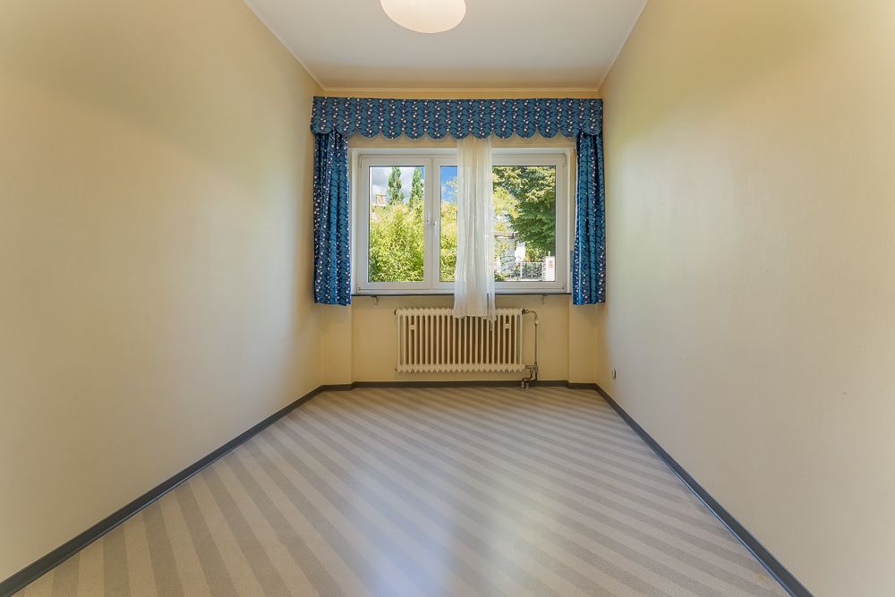 Leeres Zimmer mit gelben Wänden, gestreiftem Boden und Fenster mit blauen Vorhängen.