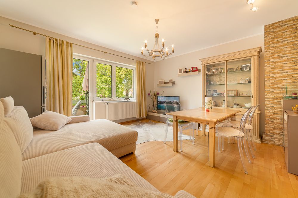 Helles Wohnzimmer mit Sofa, Esstisch, Vitrine, TV und großen Fenstern mit Ausblick ins Grüne.