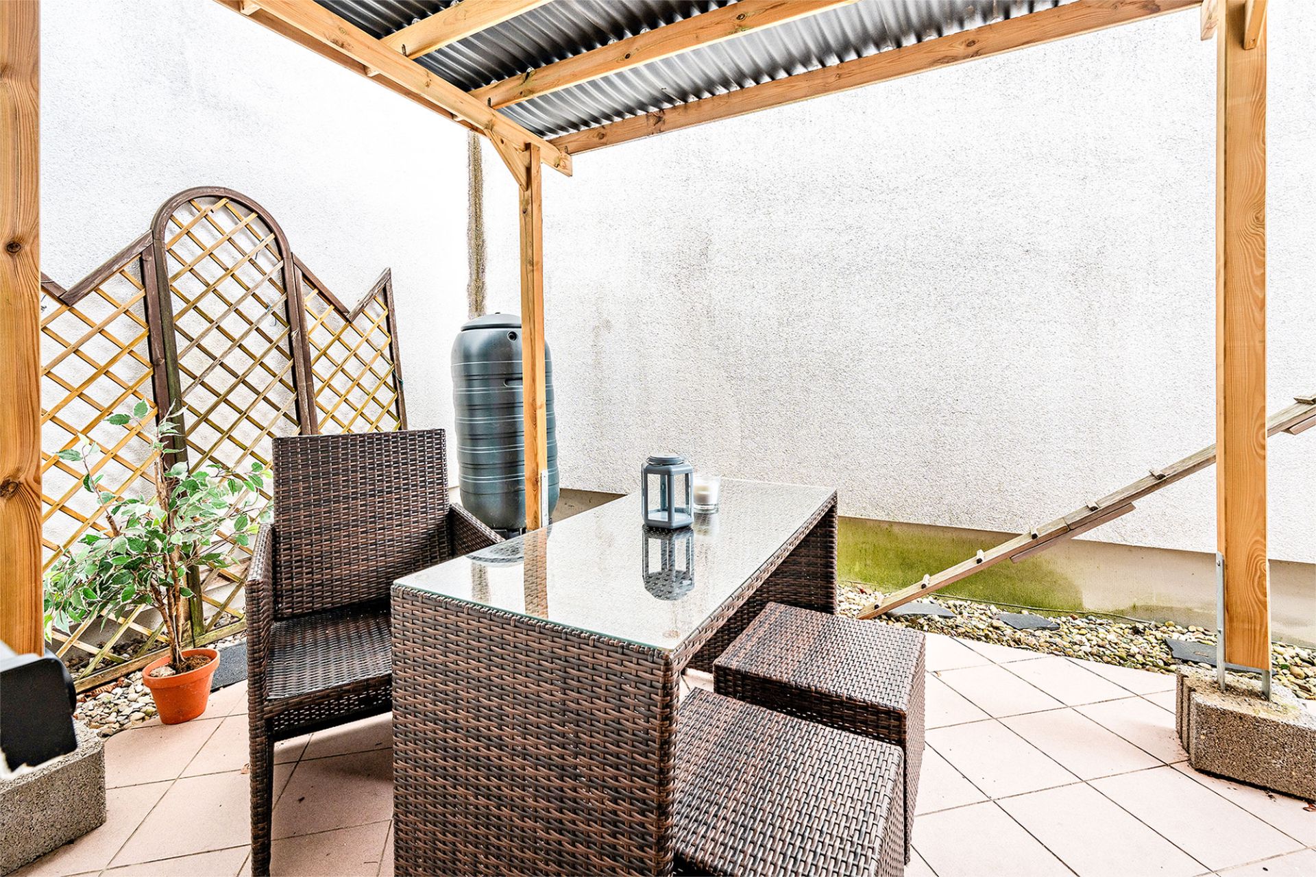 Überdachte Terrasse mit Korbmöbeln, Glastisch, Pflanze und Regentonne.