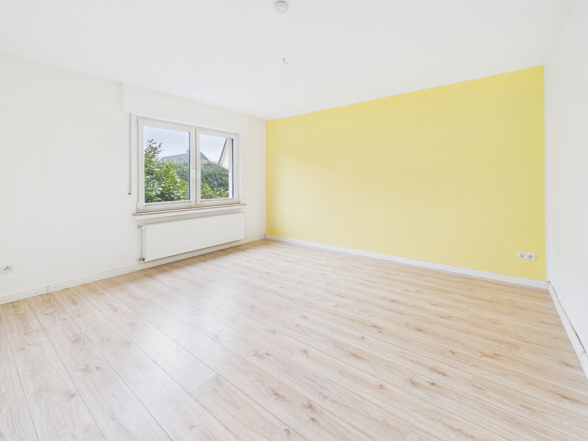 Heller leerer Raum mit gelber Wand, großem Fenster und hellem Holzfußboden.