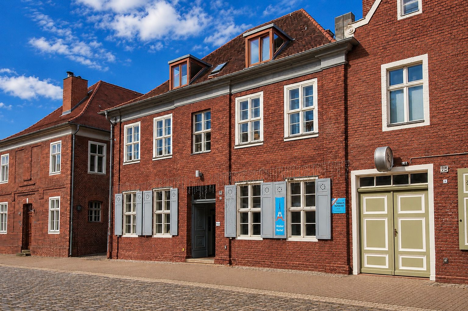 Rotes Backsteinhaus mit weißen Fenstern und zwei Eingängen an einer Kopfsteinpflasterstraße.