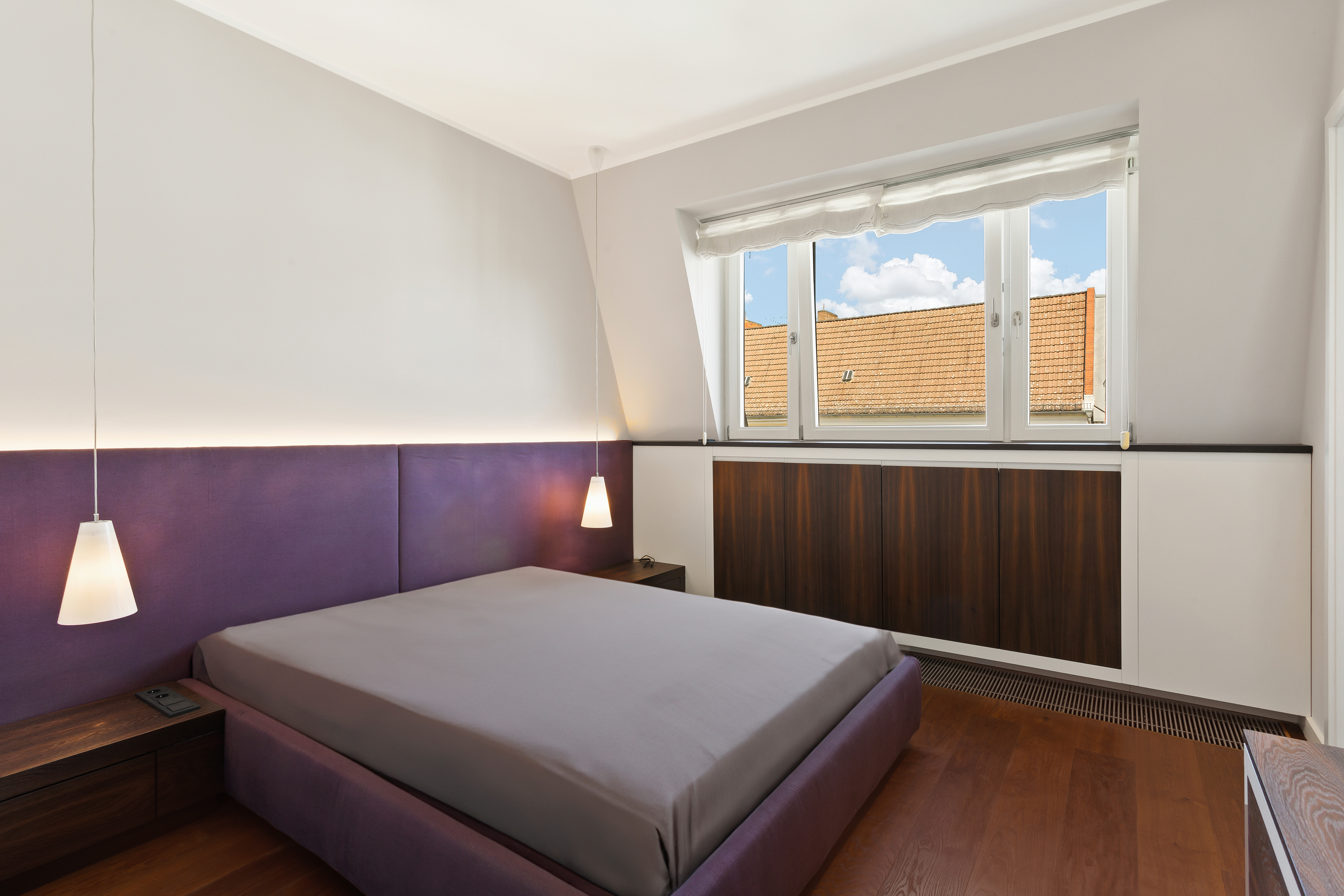 Modernes Schlafzimmer mit violettem Bett, zwei Hängelampen und großem Fenster mit Blick auf Dächer.