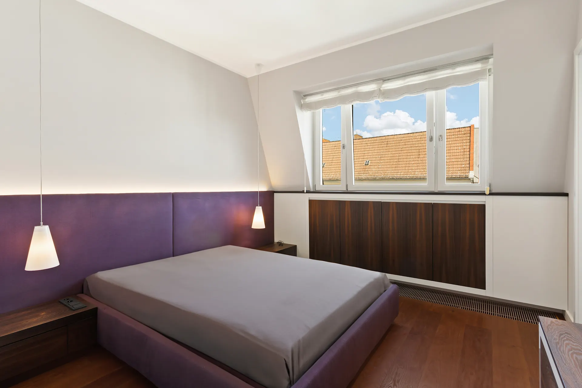 Modernes Schlafzimmer mit violettem Bett, zwei Hängelampen und großem Fenster mit Blick auf Dächer.